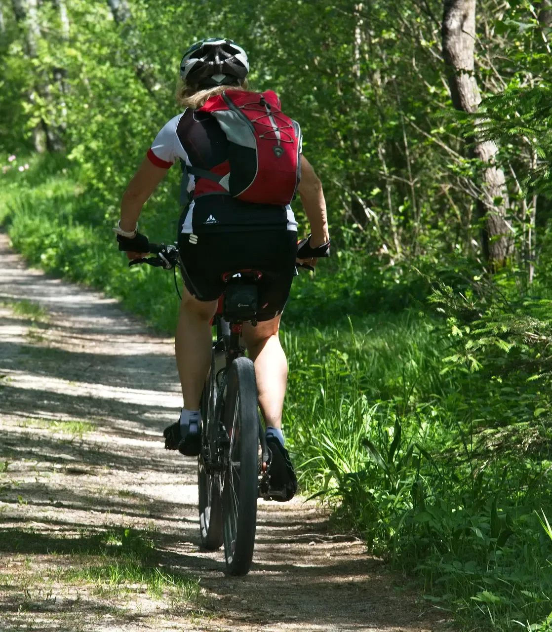A person on a bicycle is pedaling on a dirt path in the forest, wearing a helmet and a red backpack.