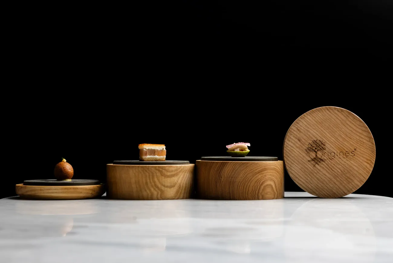 Trois petites boîtes en bois avec des couvercles, contenant des aliments, disposées sur une surface lisse et brillante devant un fond noir
