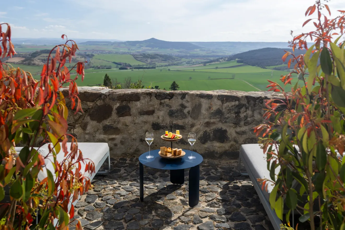 Une table avec des verres de vin et des plats est installée sur une terrasse en pierre avec vue sur une vallée verdoyante