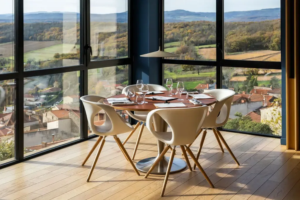 Table ronde avec quatre chaises modernes, verres et couverts, devant de grandes baies vitrées offrant une vue sur un village et des collines verdoyantes