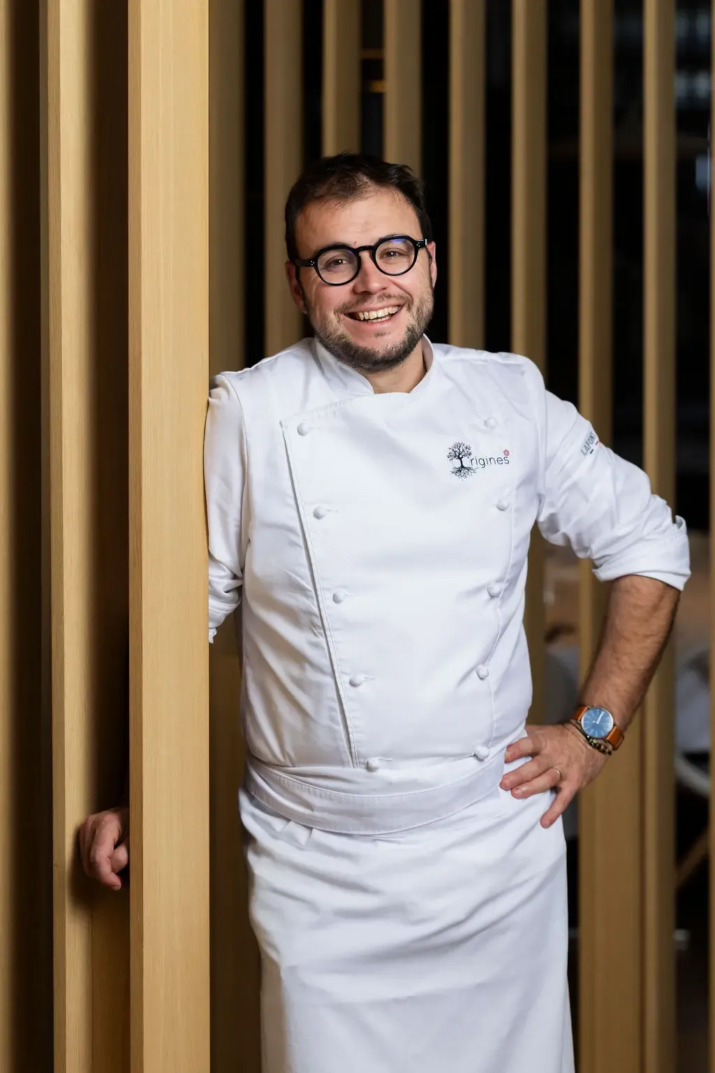 Un homme en tenue de chef de cuisine blanche sourit et pose la main sur une colonne en bois
