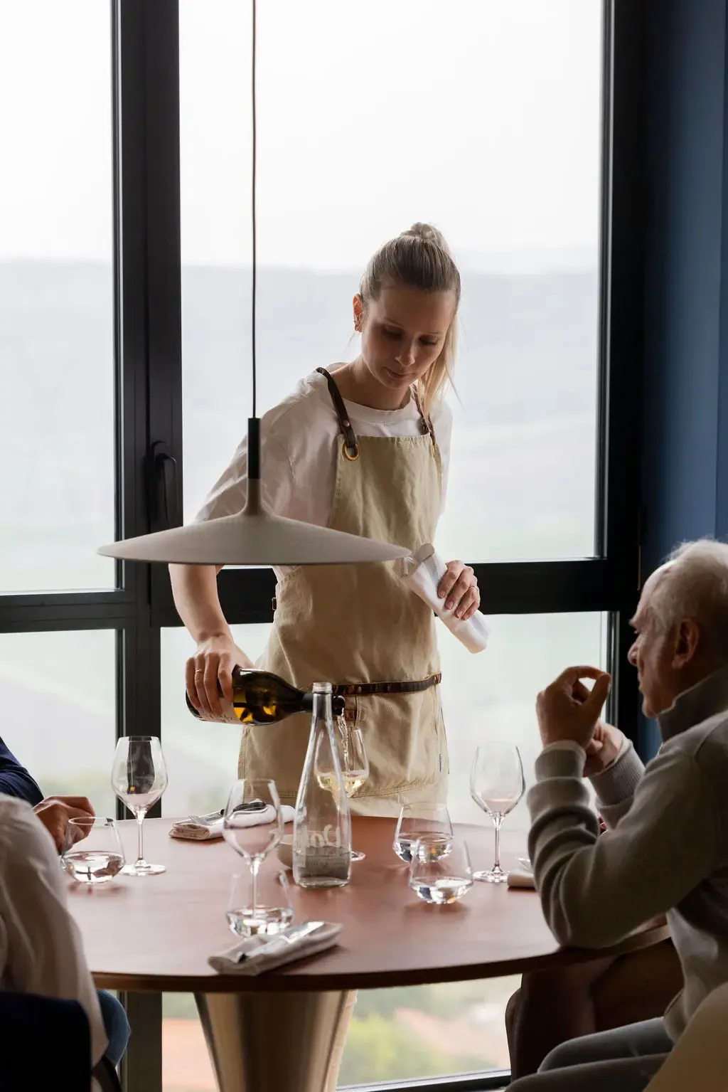 A waitress in a beige apron pours wine for a customer seated at a table with wine glasses and a water carafe.