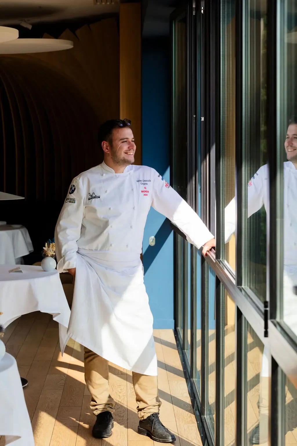Un chef en tenue blanche sourit et pose la main sur une porte vitrée dans un restaurant élégant