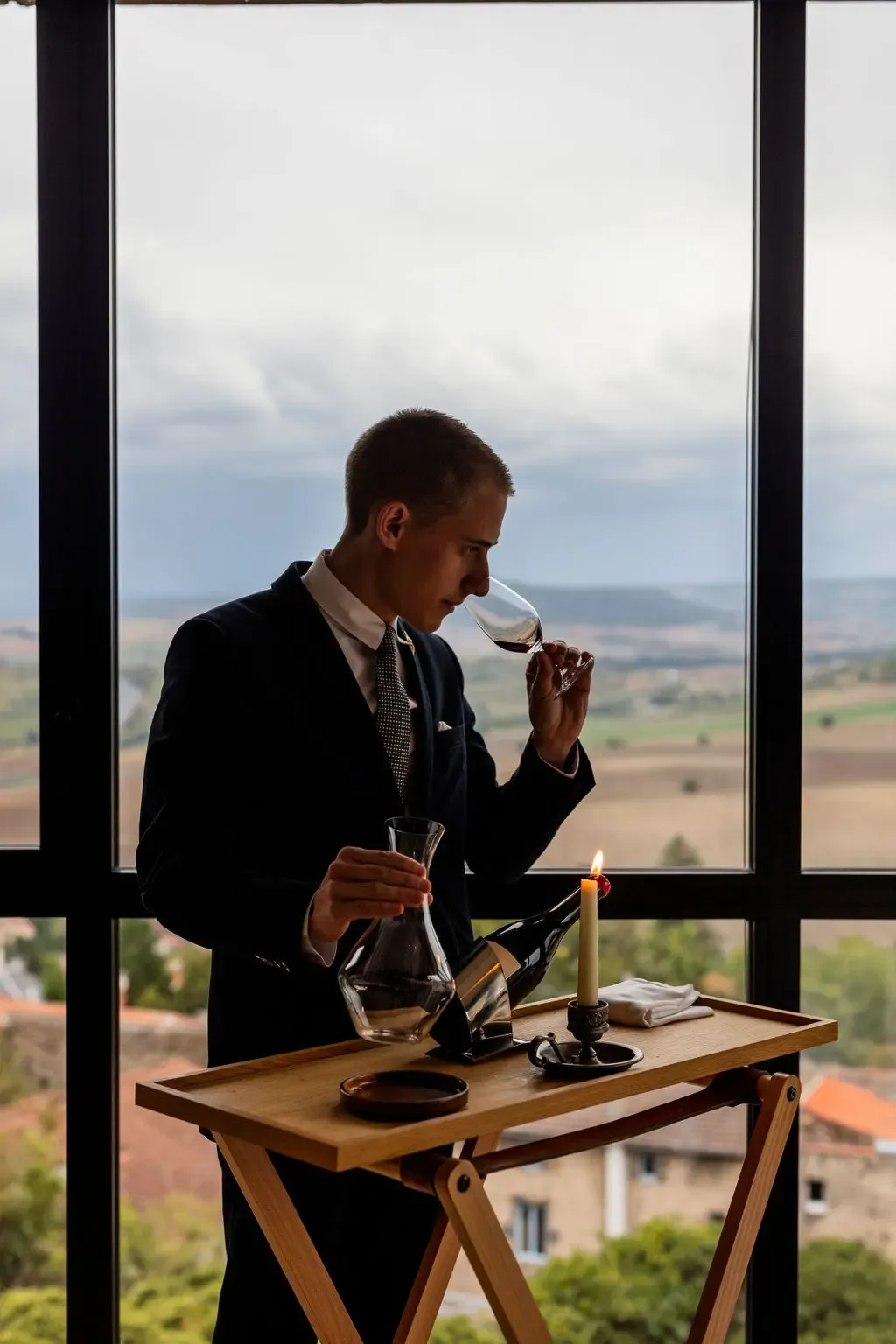 Un homme en costume tient un verre de vin et une carafe, devant une table avec une bougie et une bouteille, avec un paysage rural en arrière-plan