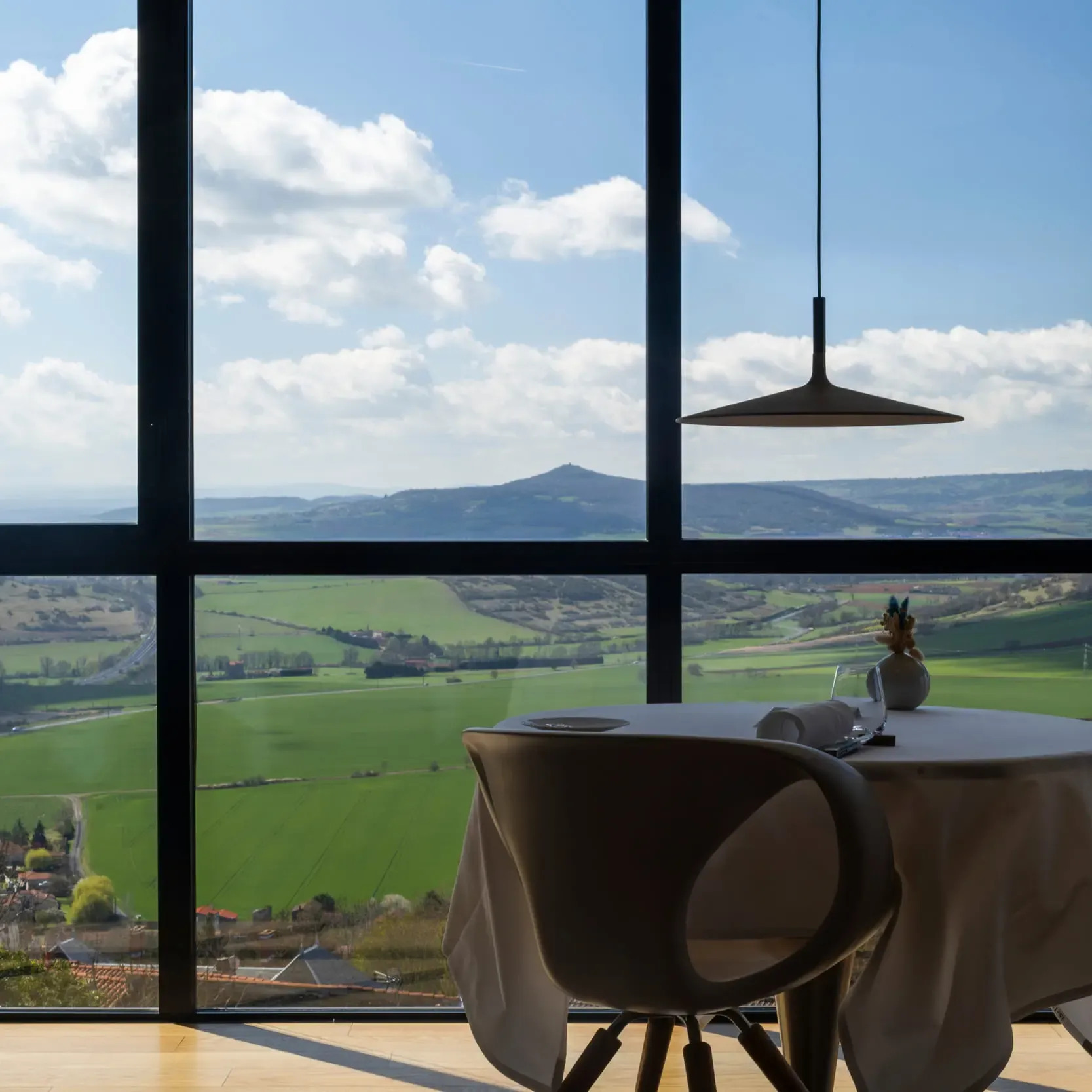 A table with a tablecloth and a vase of flowers in front of a large window offering a view of green hills and a partly cloudy sky.