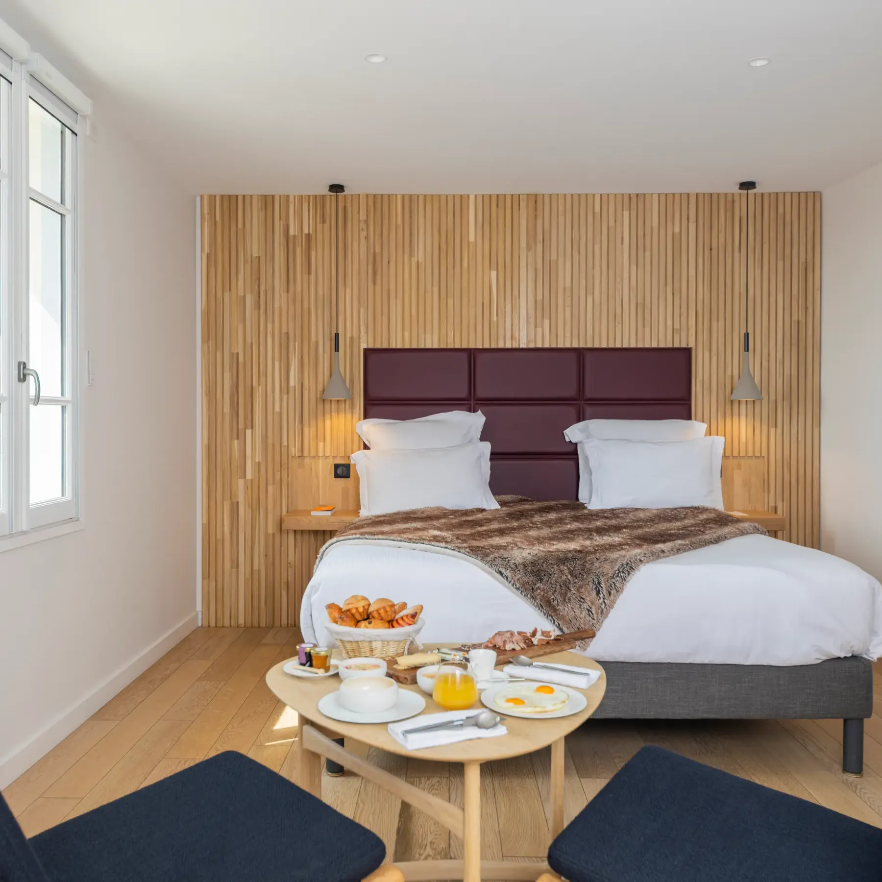 Room with double bed, wooden and velvet headboard, table with breakfast, two chairs and window