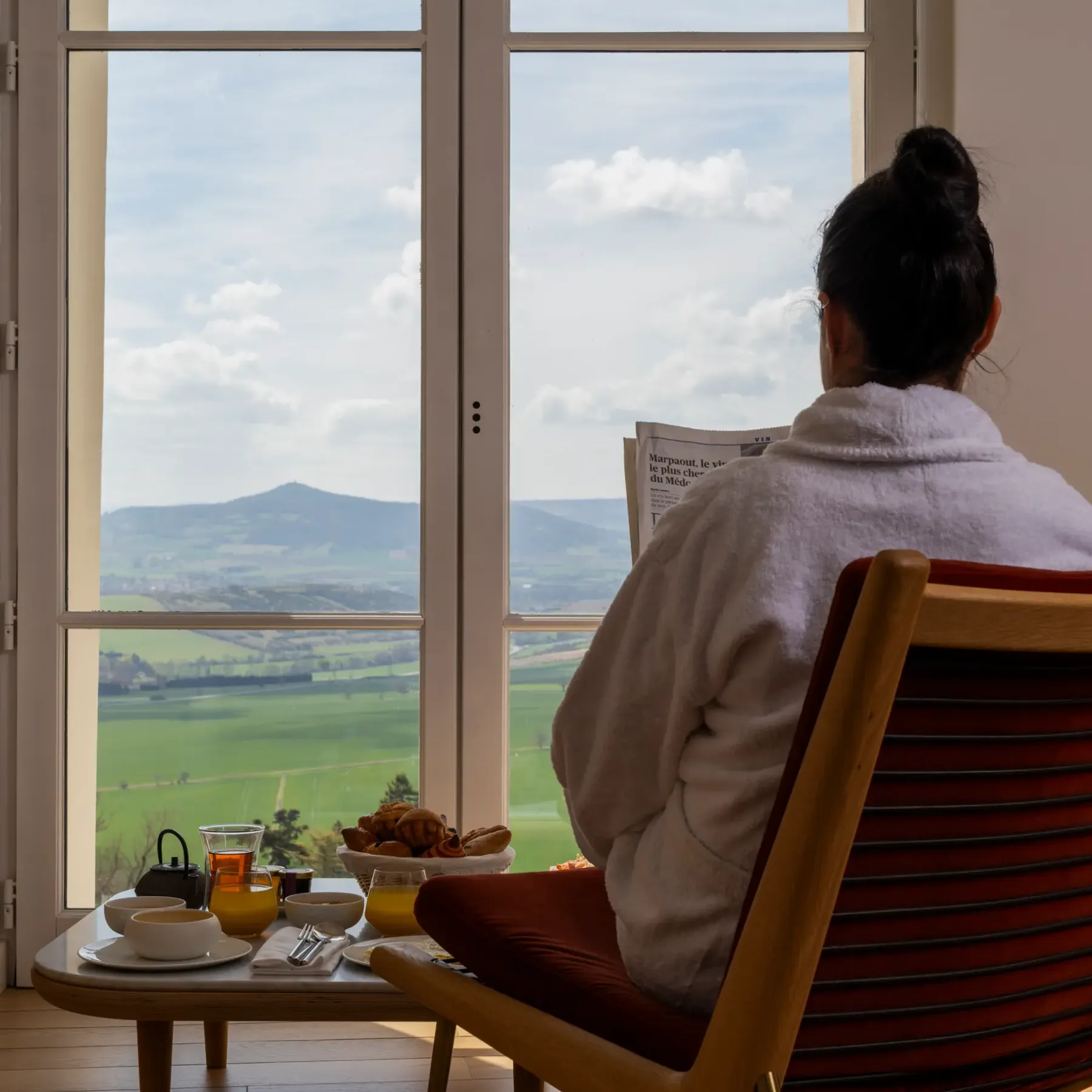 A person sitting in an armchair facing an open window, with a view of green hills, a breakfast tray beside them.
