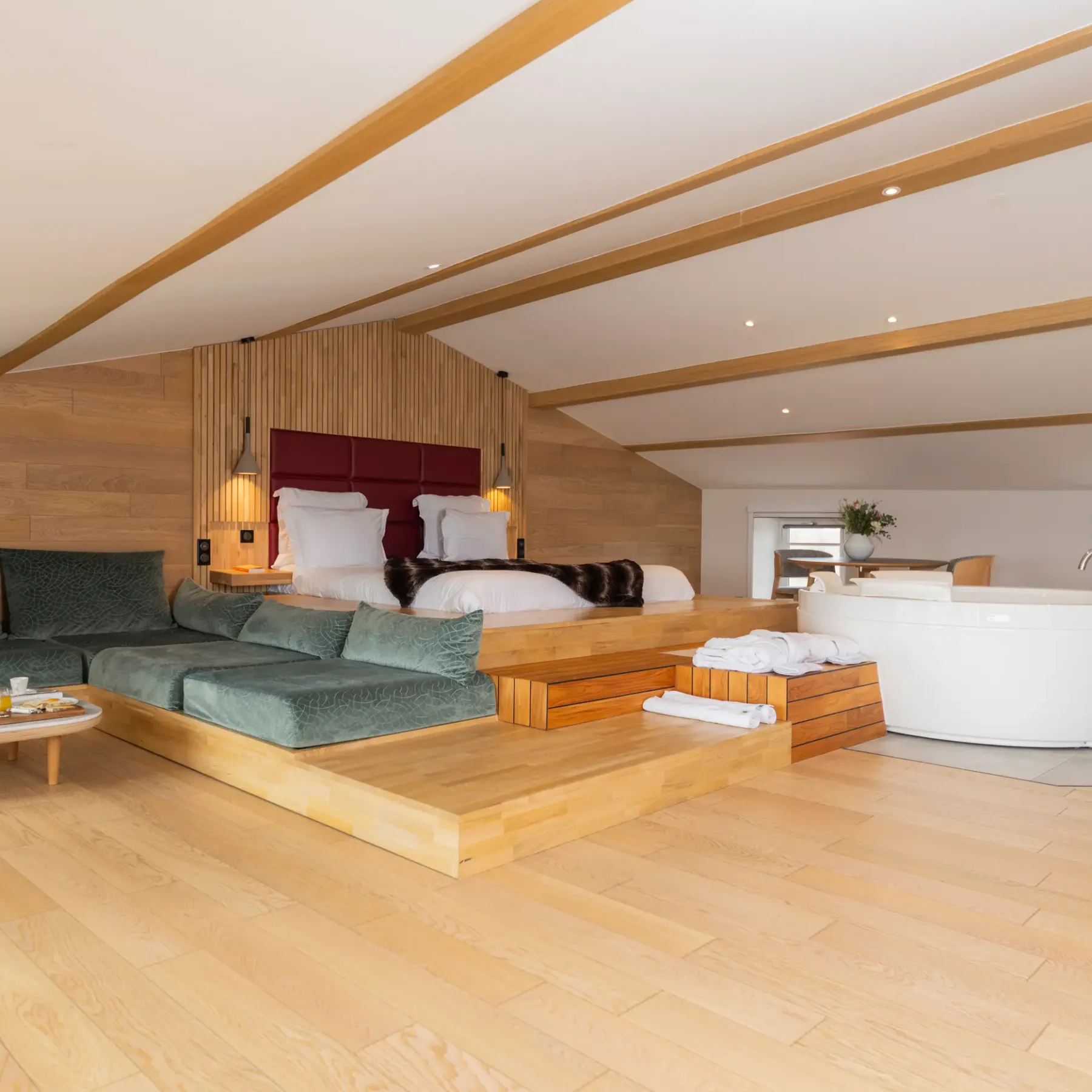 Spacious room with elevated bed, green sofa, coffee table with food, and white bathtub near a window