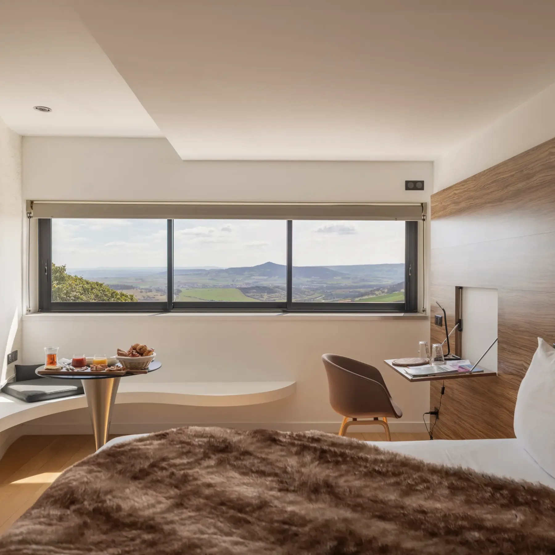 Room with bed, bedside table, chair, and desk, offering a panoramic view of a rolling landscape through a large window.
