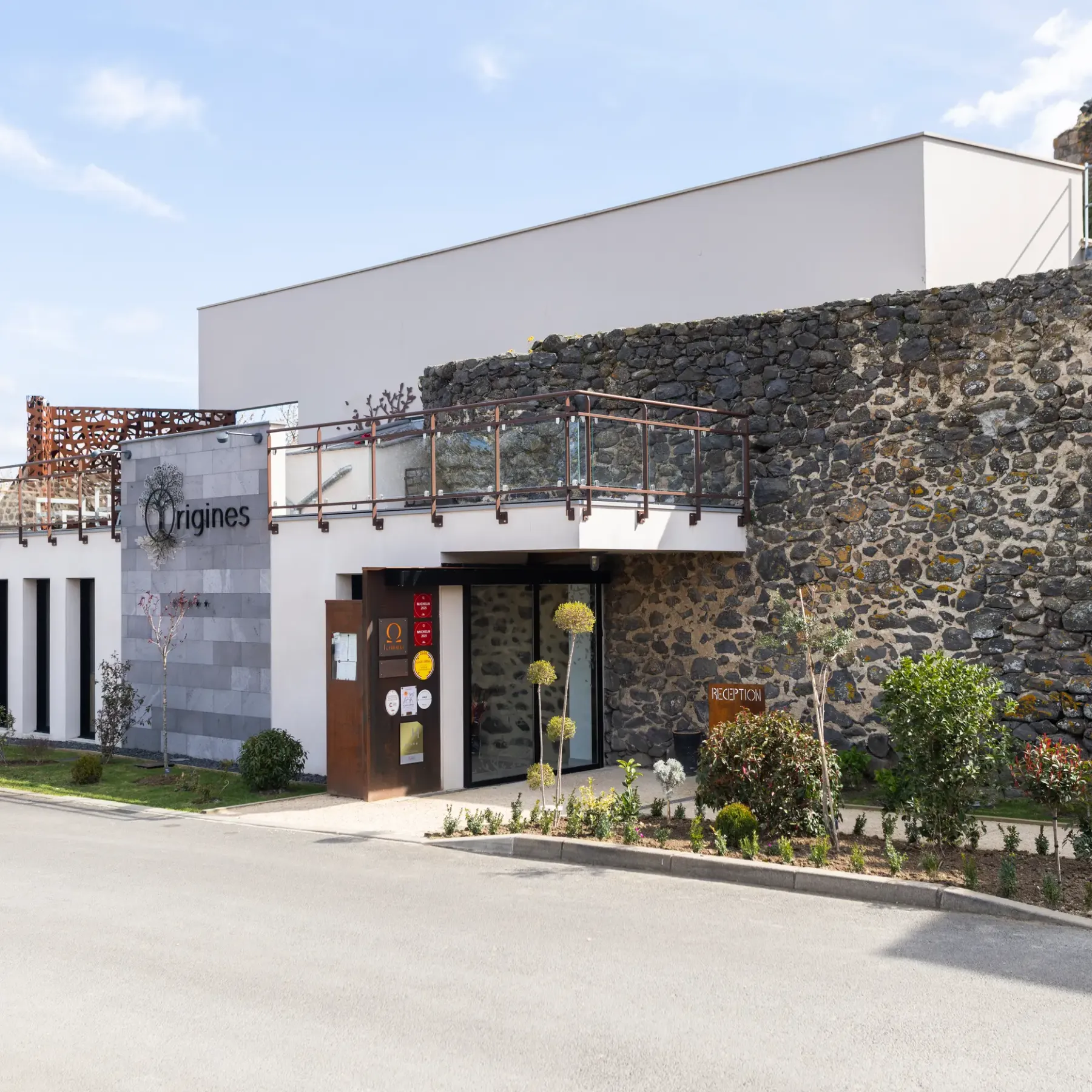 Modern building with stone facade and balconies, bearing the sign "Origines" and located in a landscaped setting