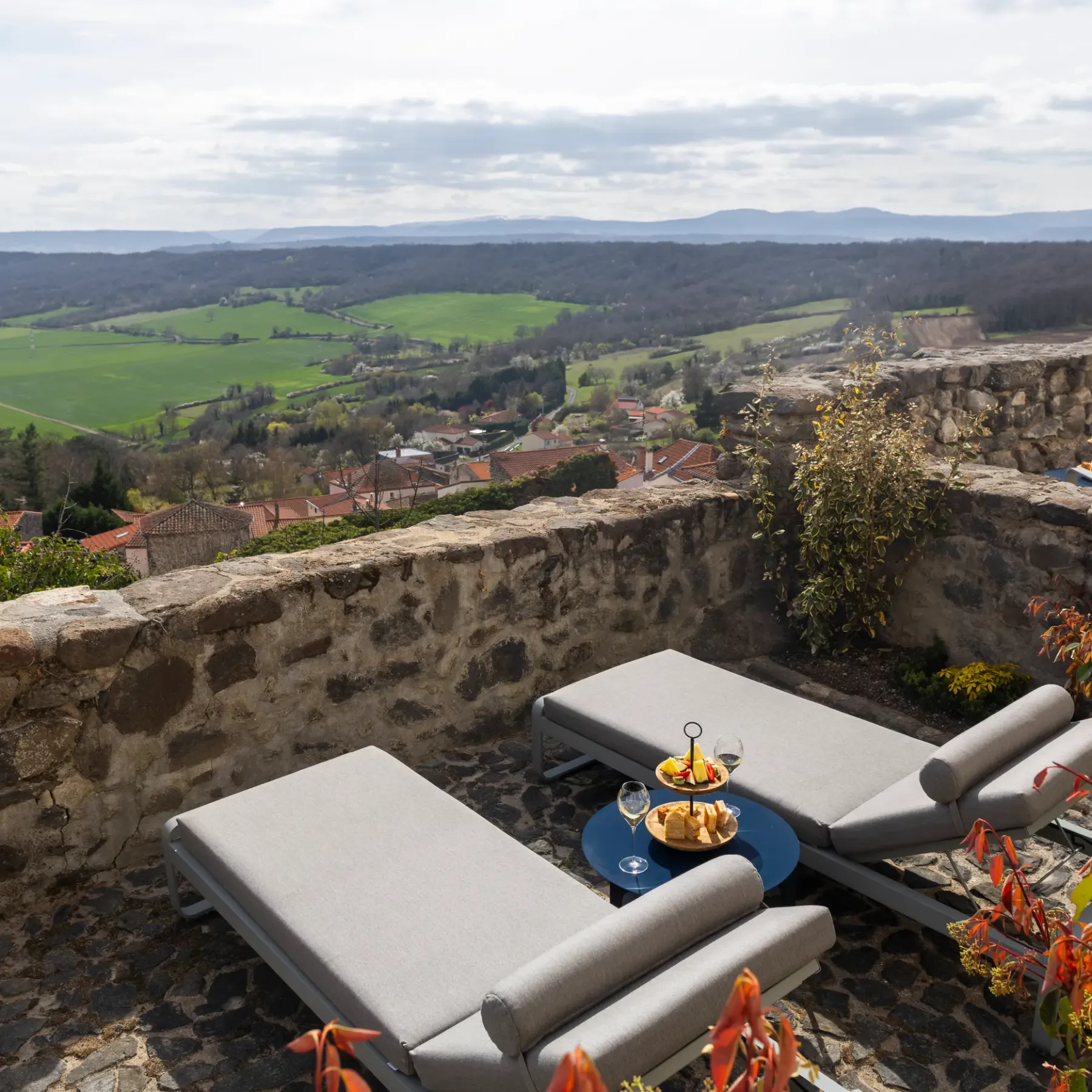 Two grey deck chairs with a low table are arranged on a stone terrace offering a view of a lush valley and distant hills.