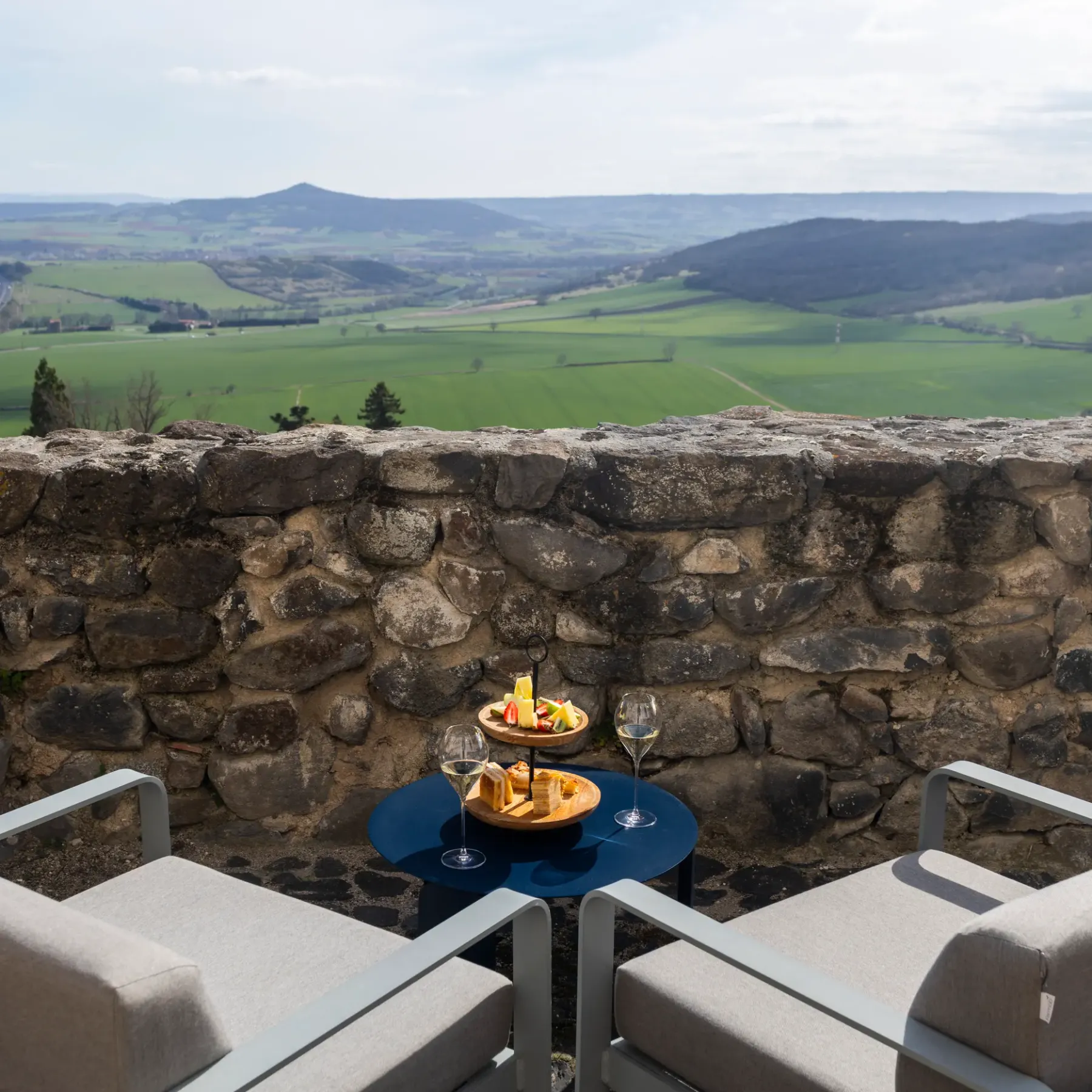 A table with glasses and a plate of food is set up in front of a stone wall, offering a view of a lush green valley.