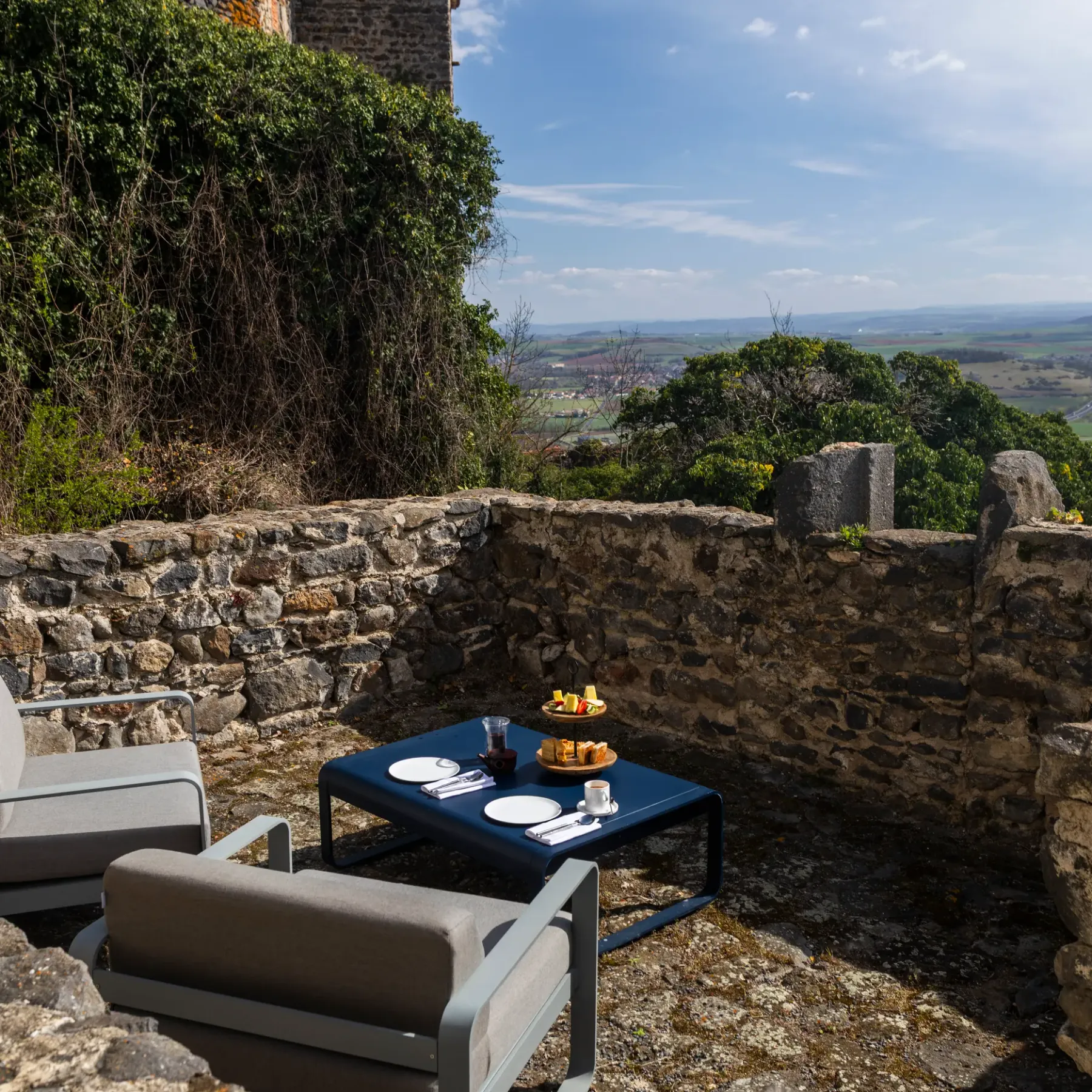An outdoor space with two deck chairs, a low table, and a breakfast tray, surrounded by stone walls and offering a view of a rolling landscape.