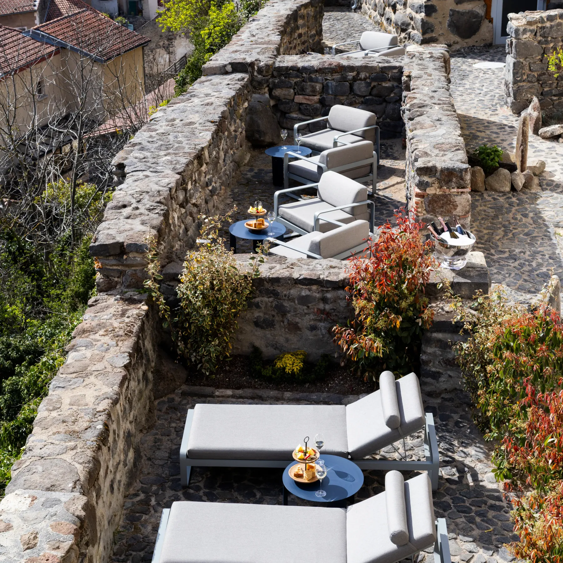 Stone terrace with sun loungers and low tables, overlooking a village with red-tiled roofs and stone walls.