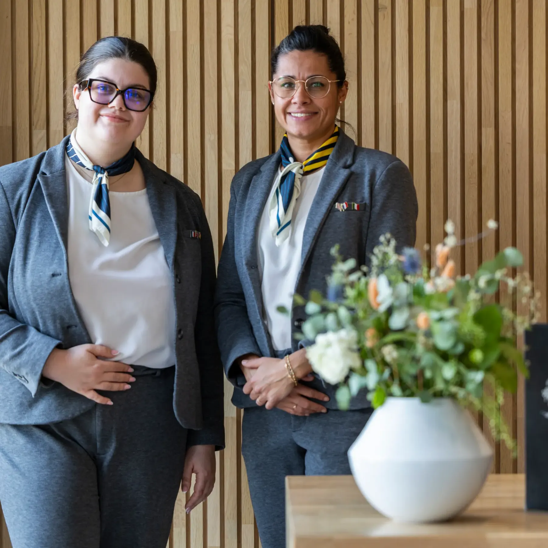 Two women in suits with scarves and badges stand in front of a wooden wall, with a vase of flowers and a laptop on a table beside them.
