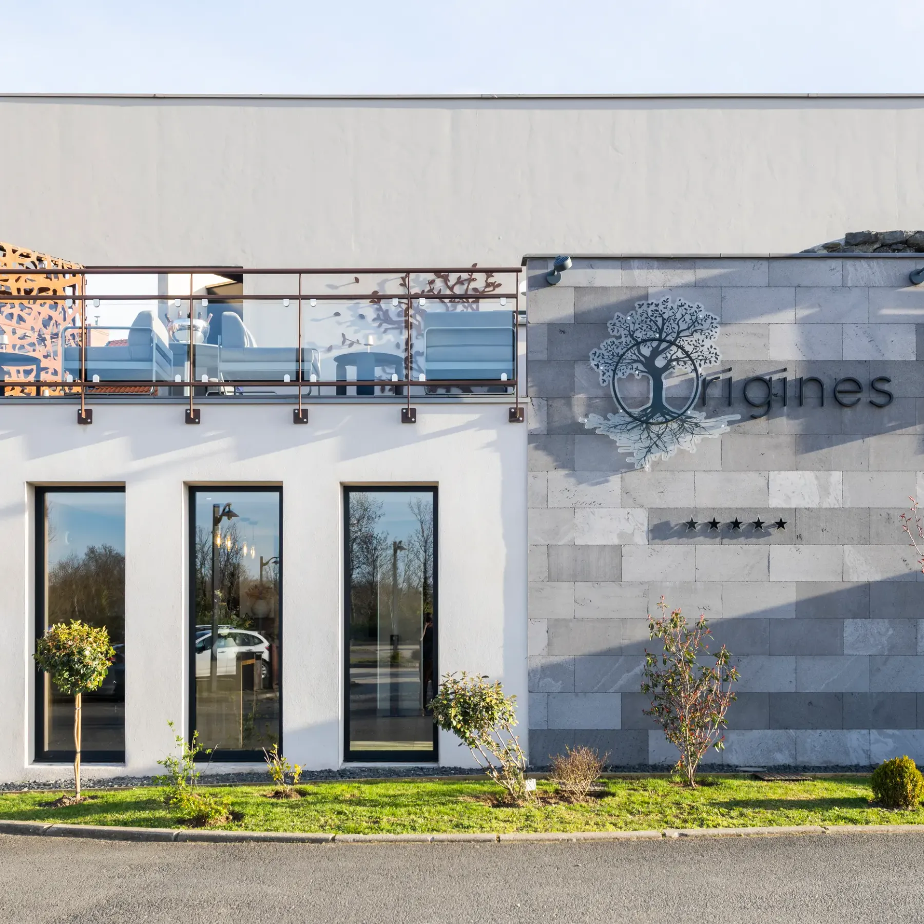 Modern building with white and grey facade, metal and vegetated balcony railing, and "Origines" sign with a tree logo