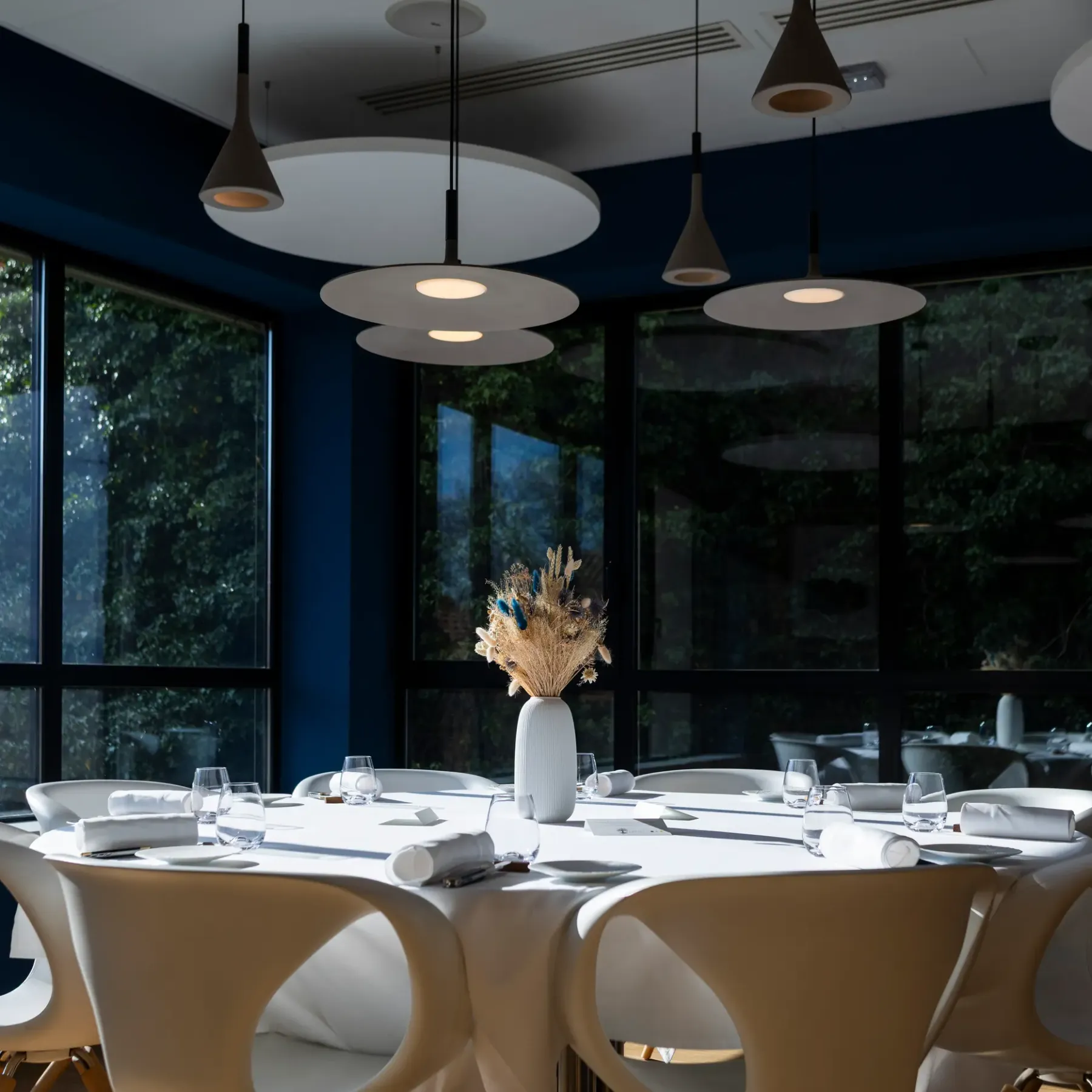 Table set with glasses and napkins in a modern dining room with large windows, lit by circular pendant lights.