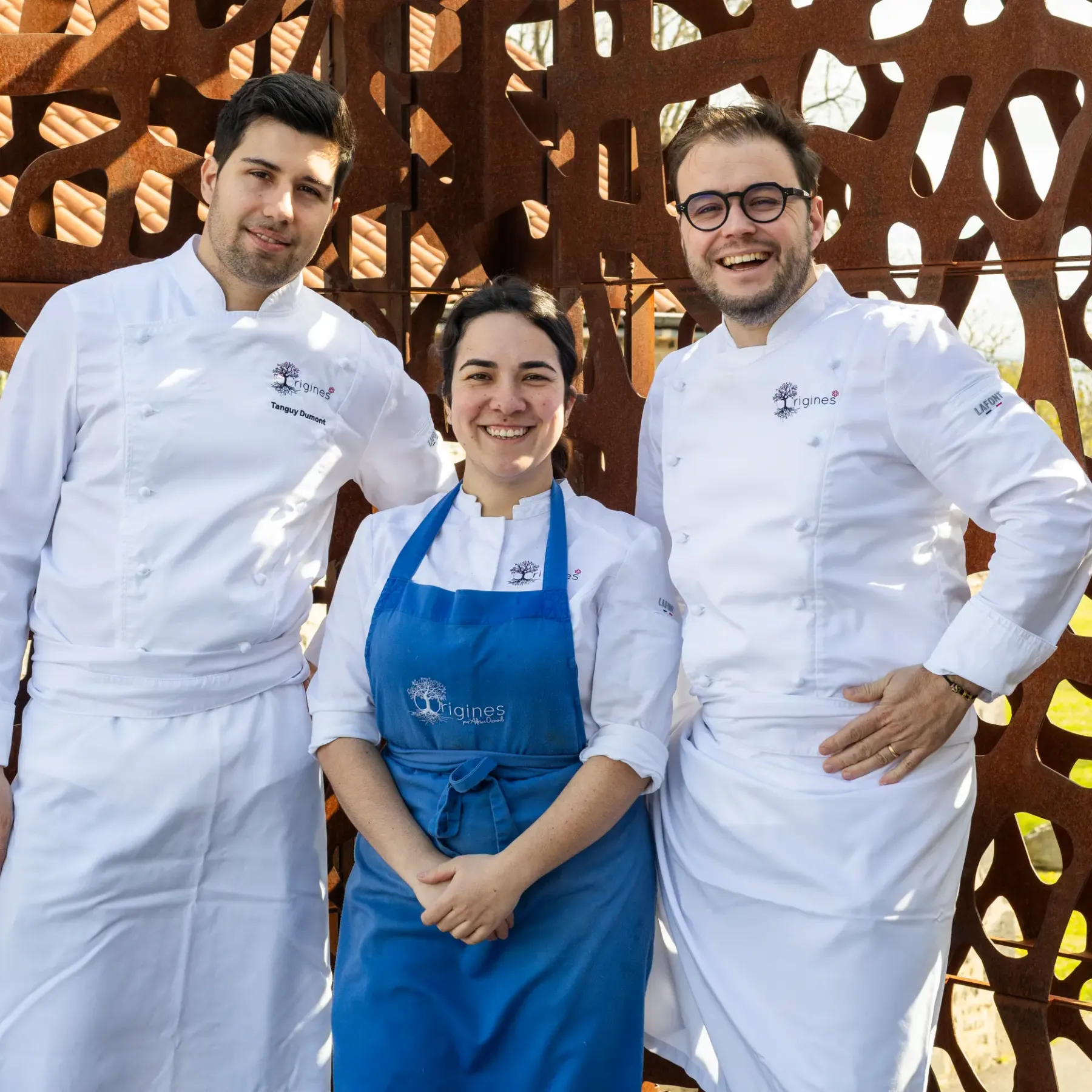 Three people in professional kitchen attire pose in front of an openwork metal structure outdoors.