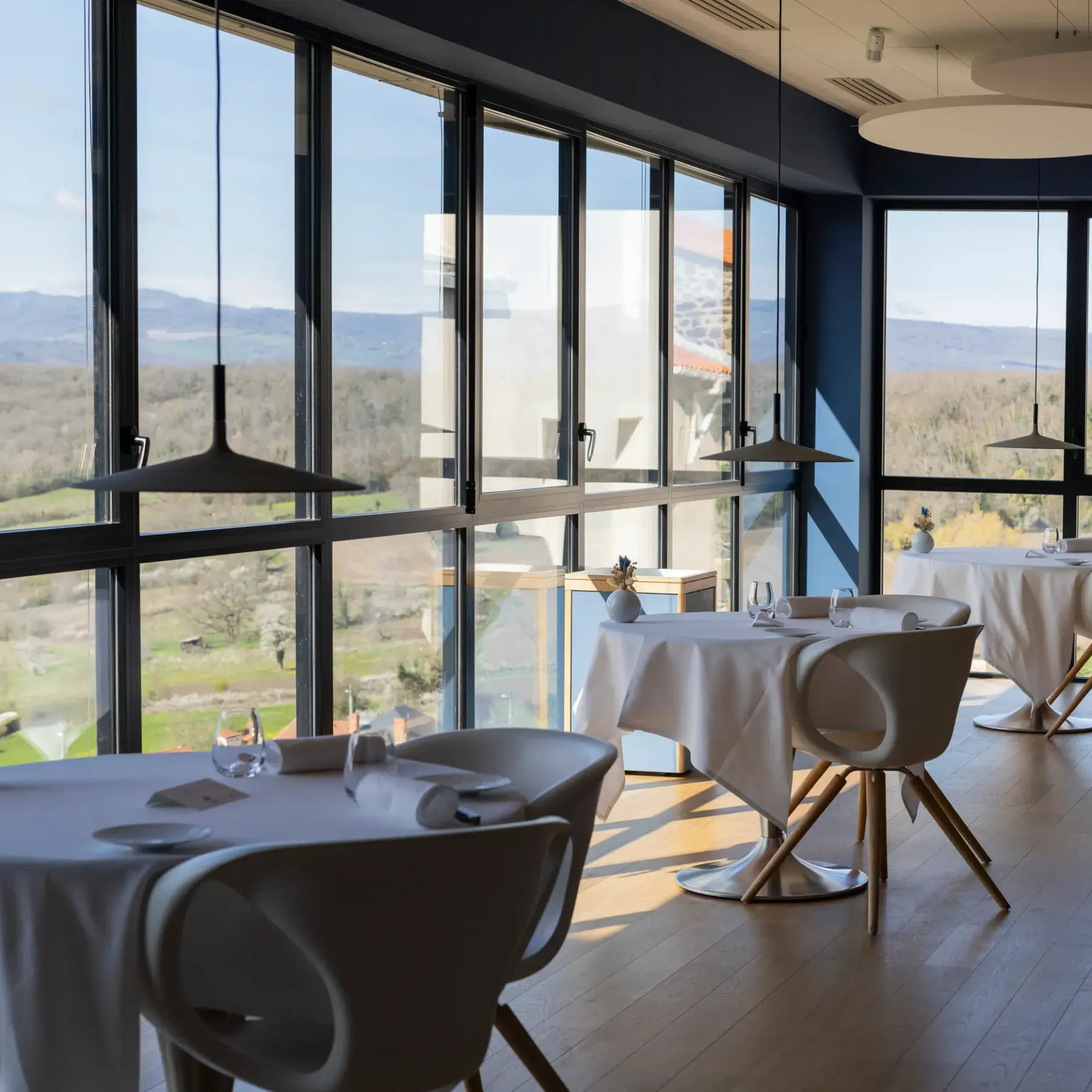 Restaurant room with modern tables and chairs, panoramic view of a hilly landscape through large glass windows.