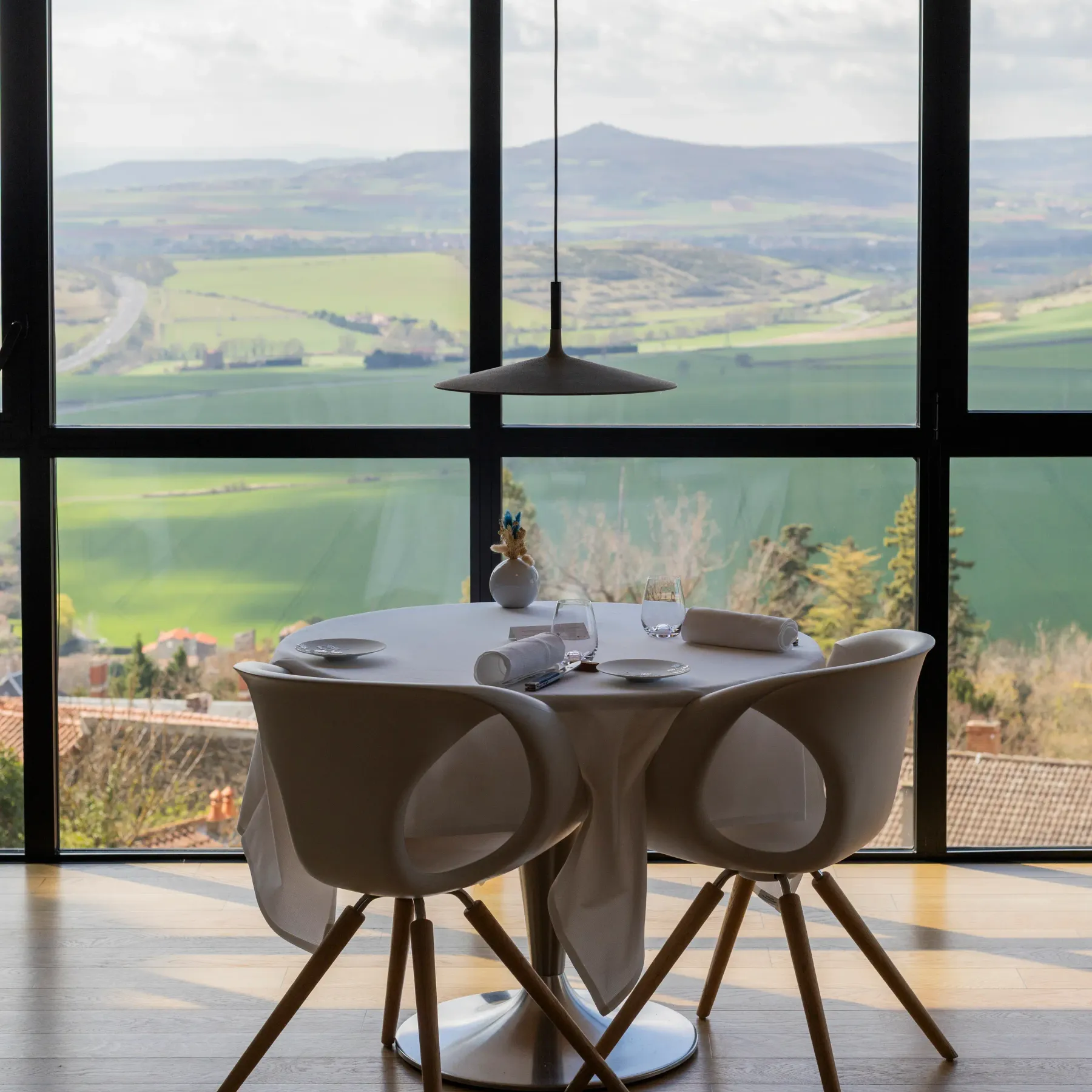 Round table with two chairs in front of a large window offering a view of a hilly landscape