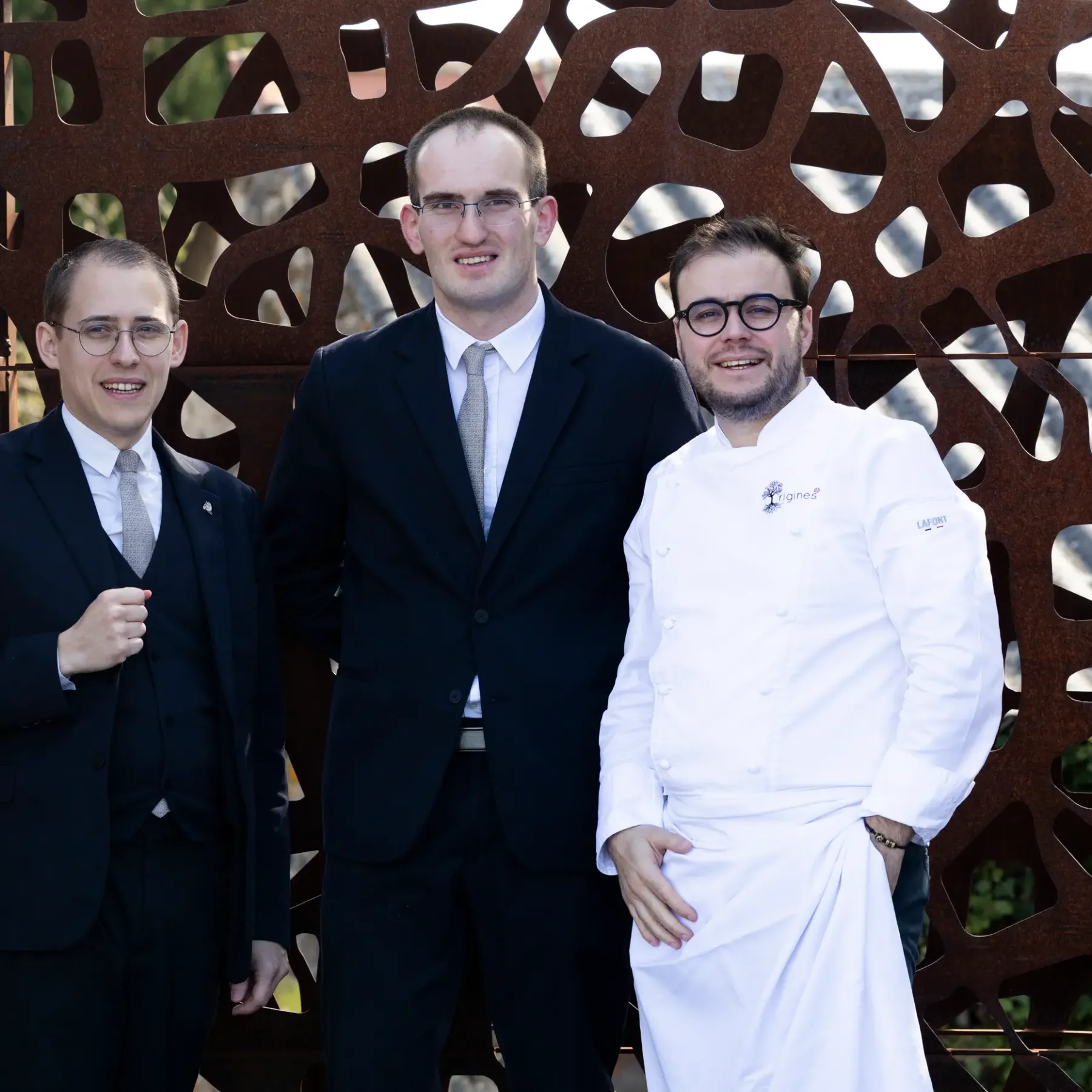 Three men in suits and white jackets pose in front of a perforated metal structure.