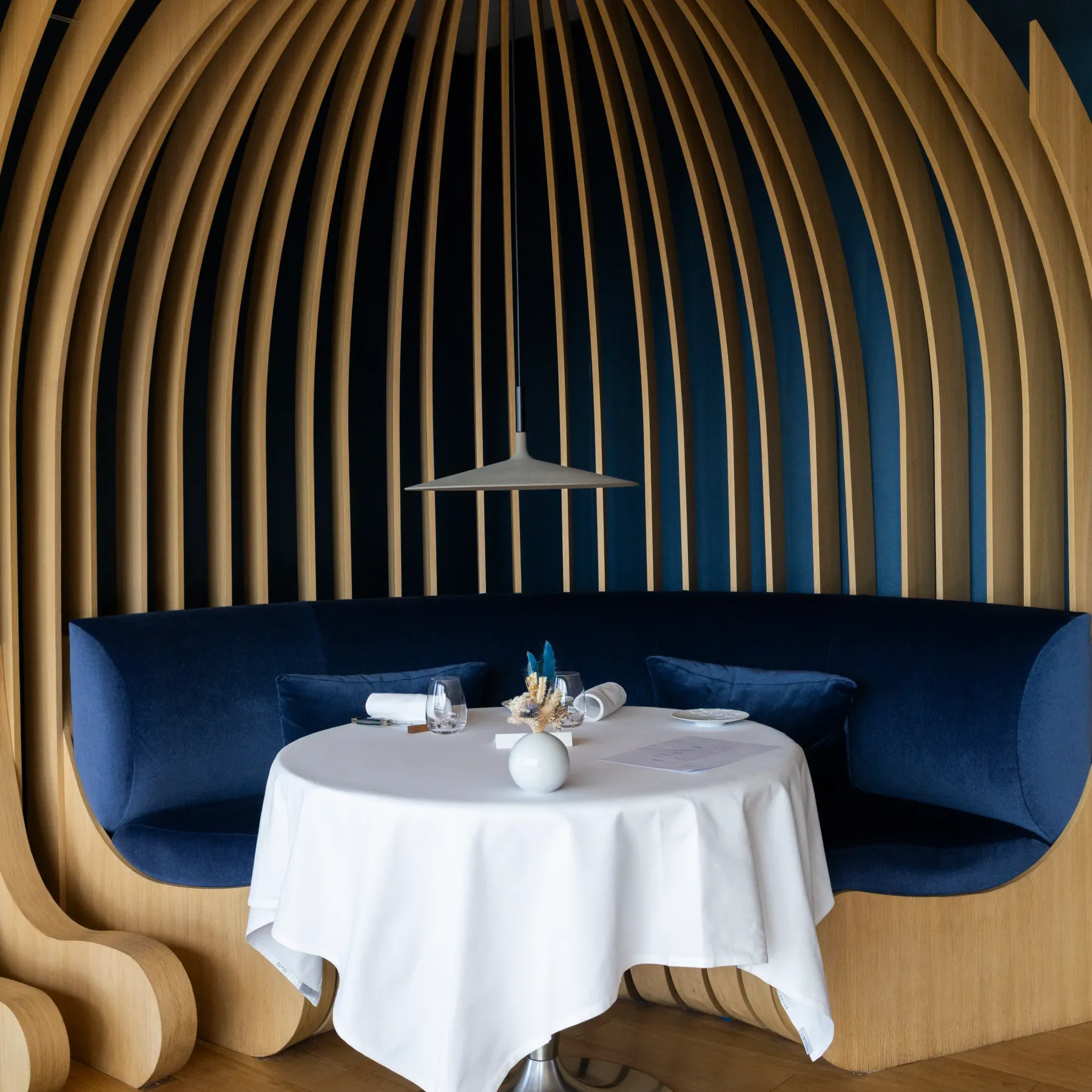 An interior space with a round table covered by a white tablecloth, surrounded by blue benches, under a wooden structure and suspended lighting.
