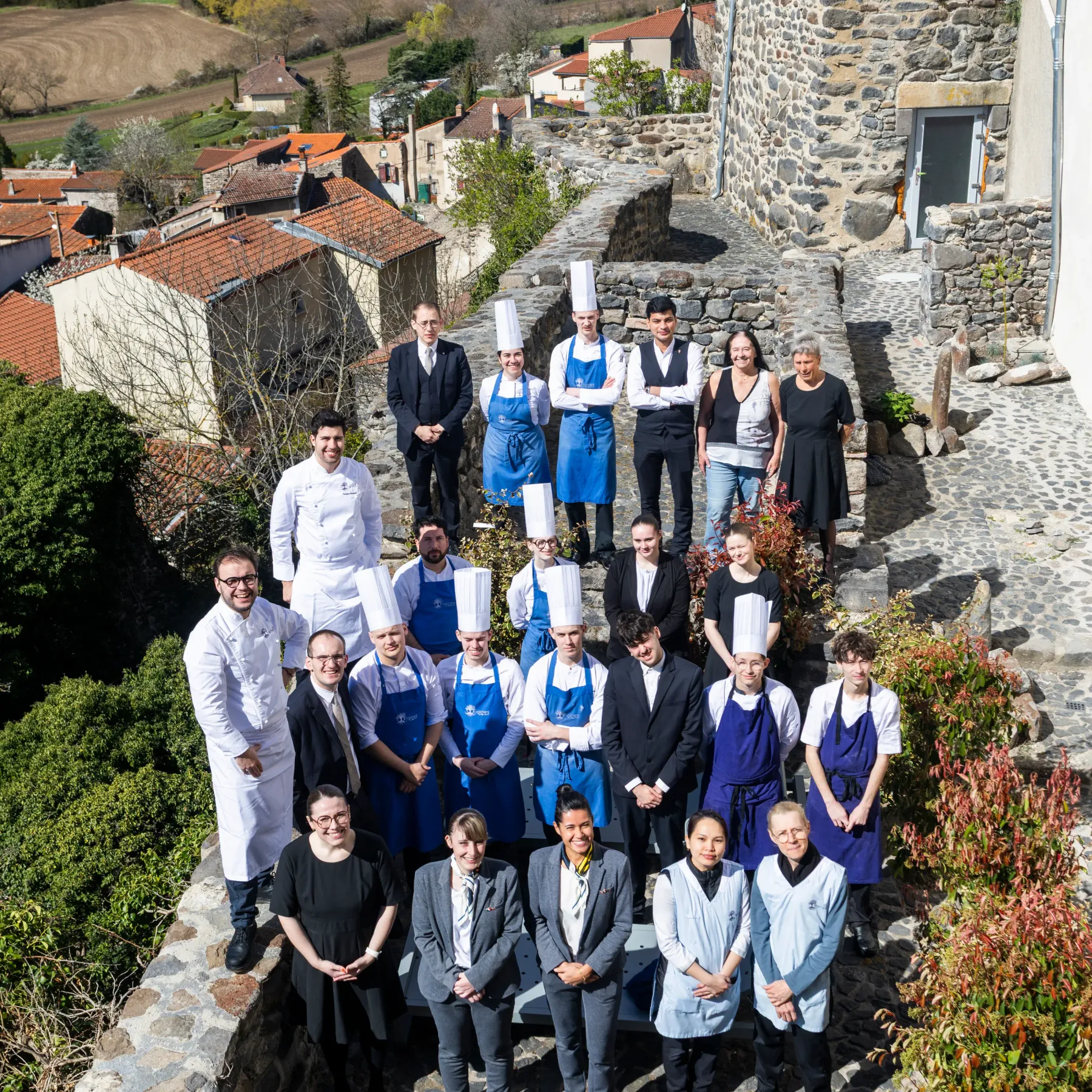 A group of people in kitchen attire and formal clothing pose in front of a stone building in a village with a view of the countryside.