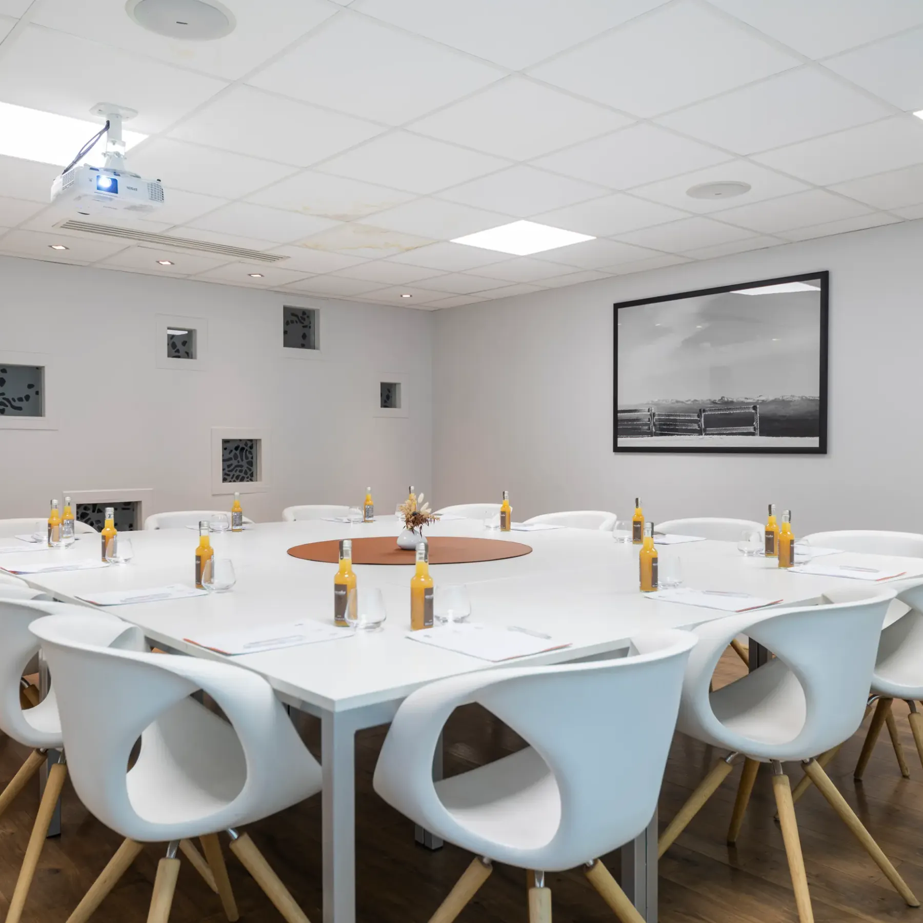 Meeting room with a white round table surrounded by chairs, bottles and glasses on the table, a projector on the ceiling and artworks on the walls.