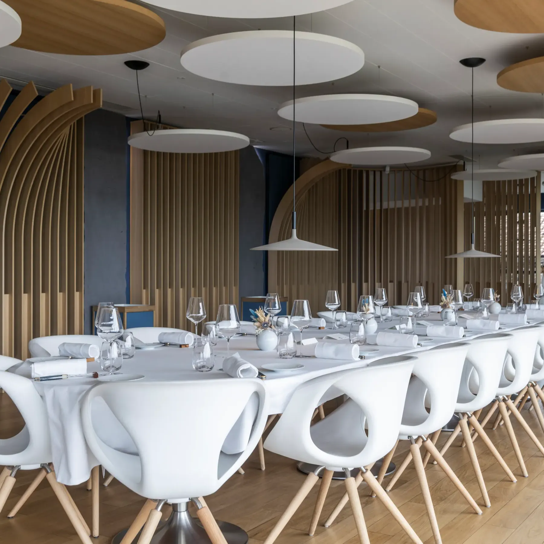 A long rectangular table with glasses, cutlery, and napkins, surrounded by white chairs, in a modern dining room with wooden walls and circular pendant lights.