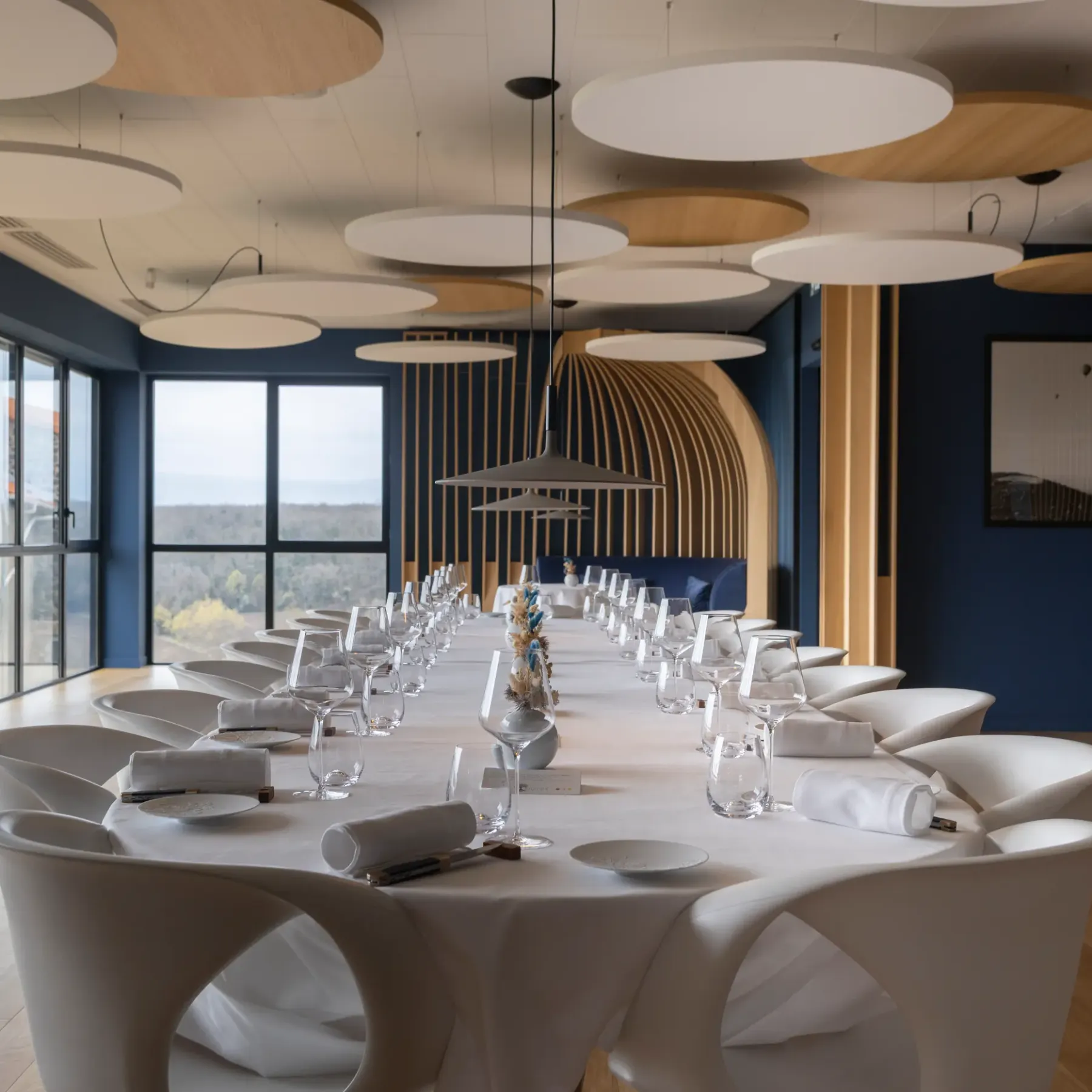 A long meeting table with modern chairs, glasses, and napkins, in a room with large windows and circular pendant lights.