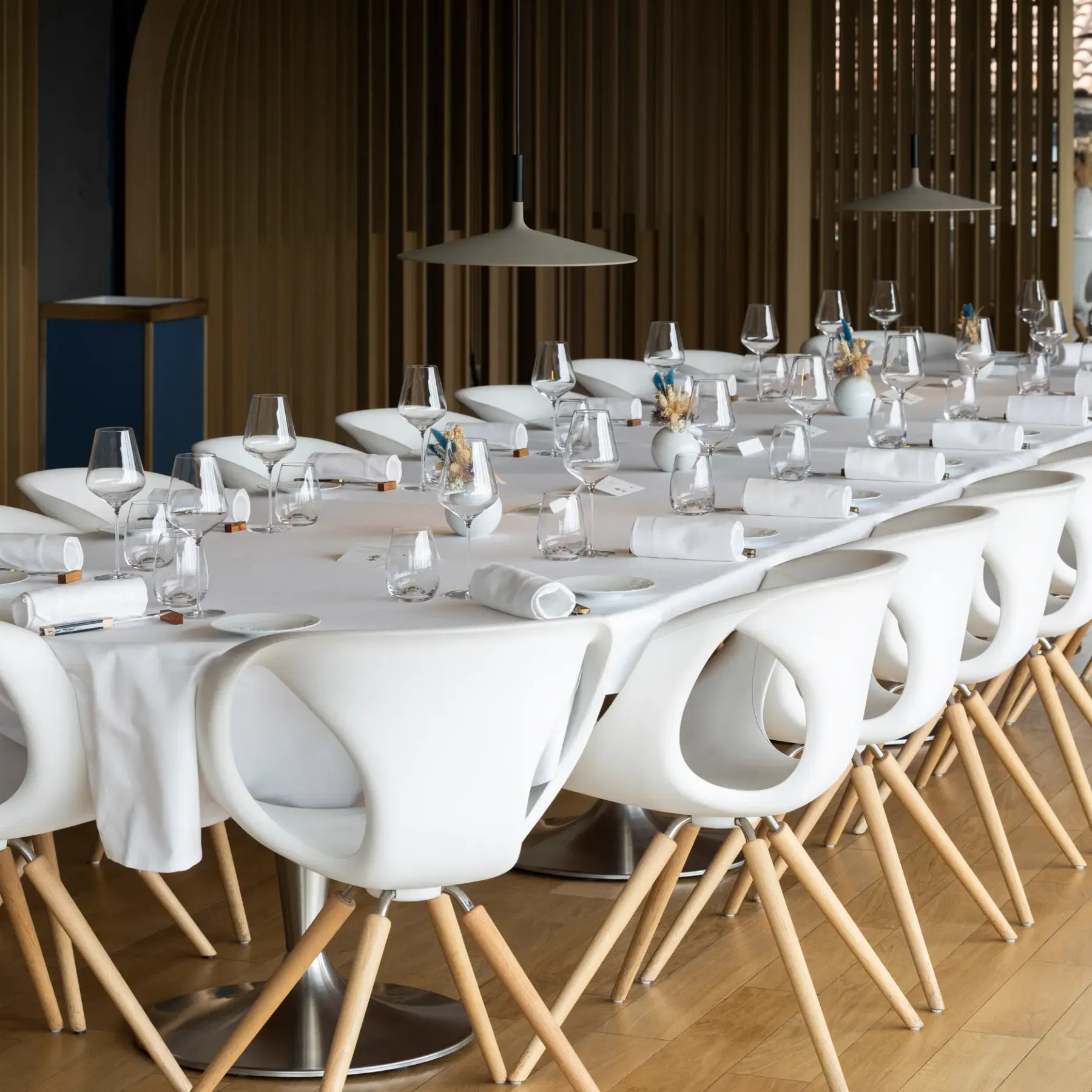 A long rectangular table is set with glasses, napkins, and small vases containing flowers, surrounded by white chairs with wooden legs, in a dining room with large windows.