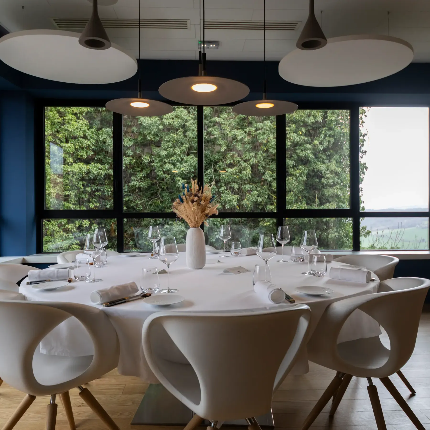 A round table with glasses and cutlery, surrounded by chairs, in a room with large windows offering a view of nature.