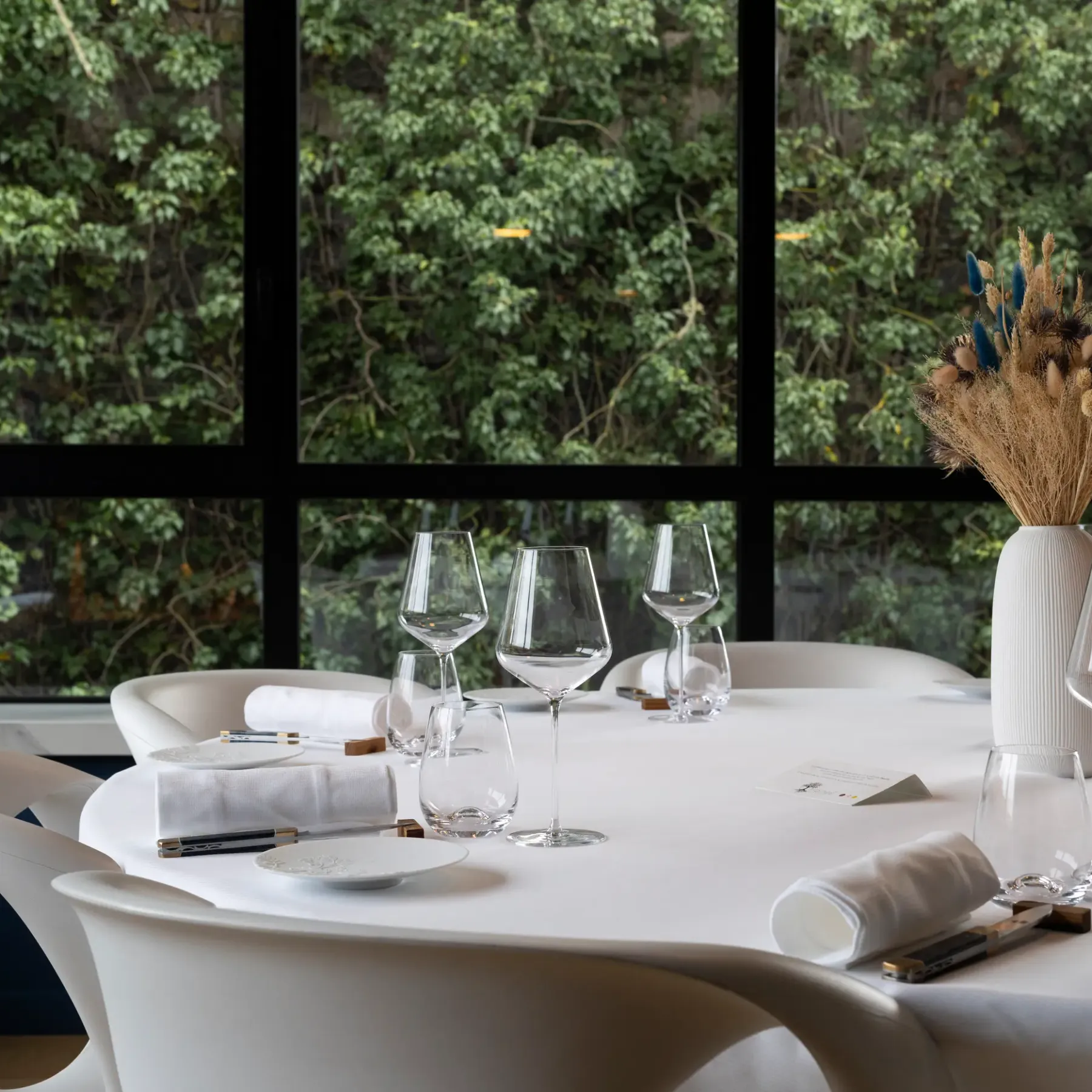 Table set with wine glasses, napkins, and a bouquet of dried flowers in front of a large window overlooking a forest.