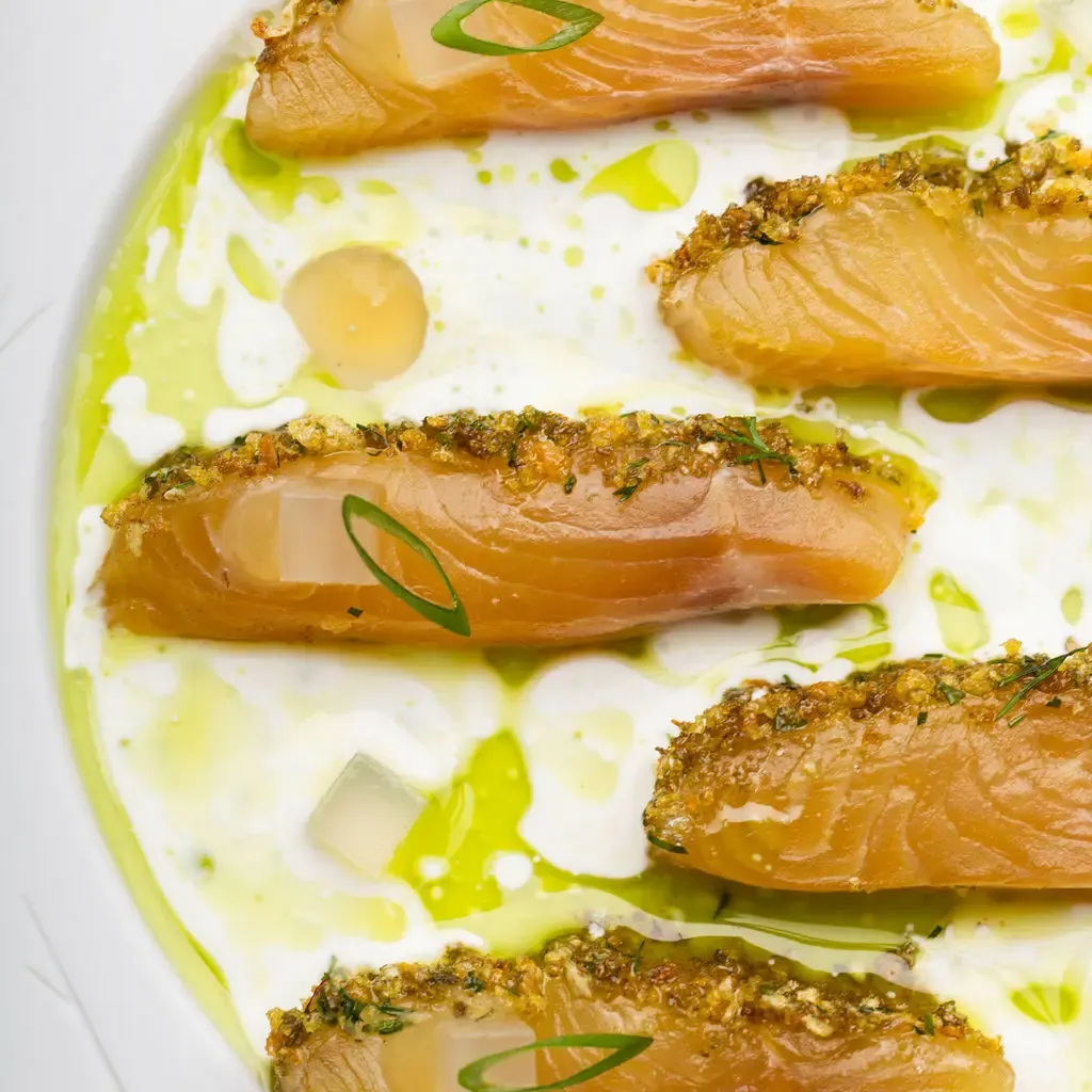 Four fish fillets garnished with herbs and panko, arranged on a green and white sauce in a white plate