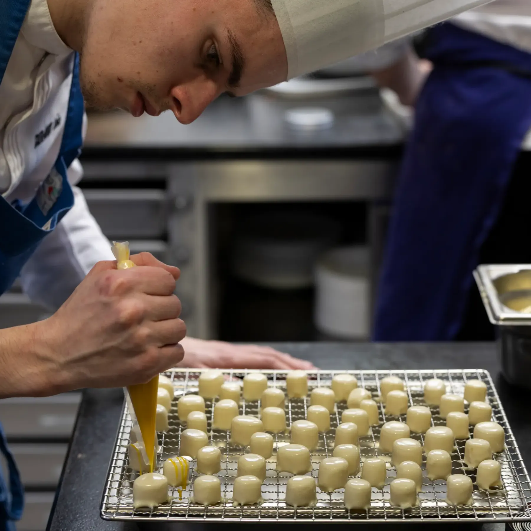 A chef in uniform pours a yellow mixture onto small squares of dough arranged on a metal grid.