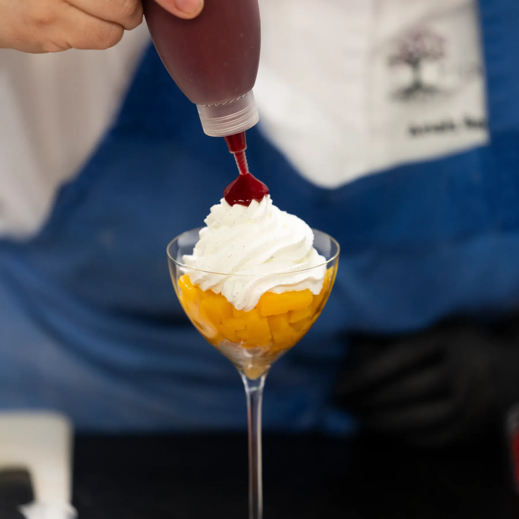 A person pours a red sauce over a dessert cup containing fruit and whipped cream.