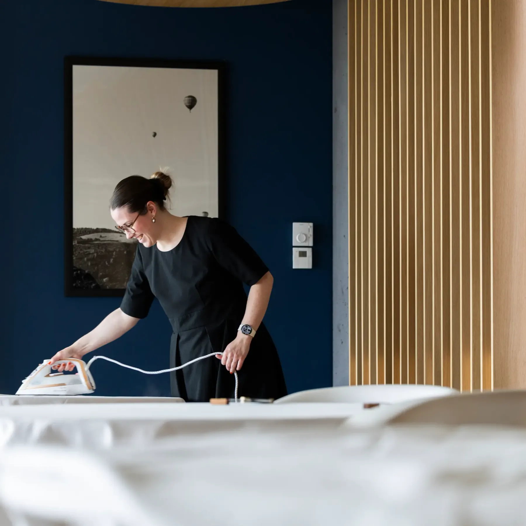 A woman is using an iron on a table covered with a white cloth in a room with dark blue walls and beige curtains.