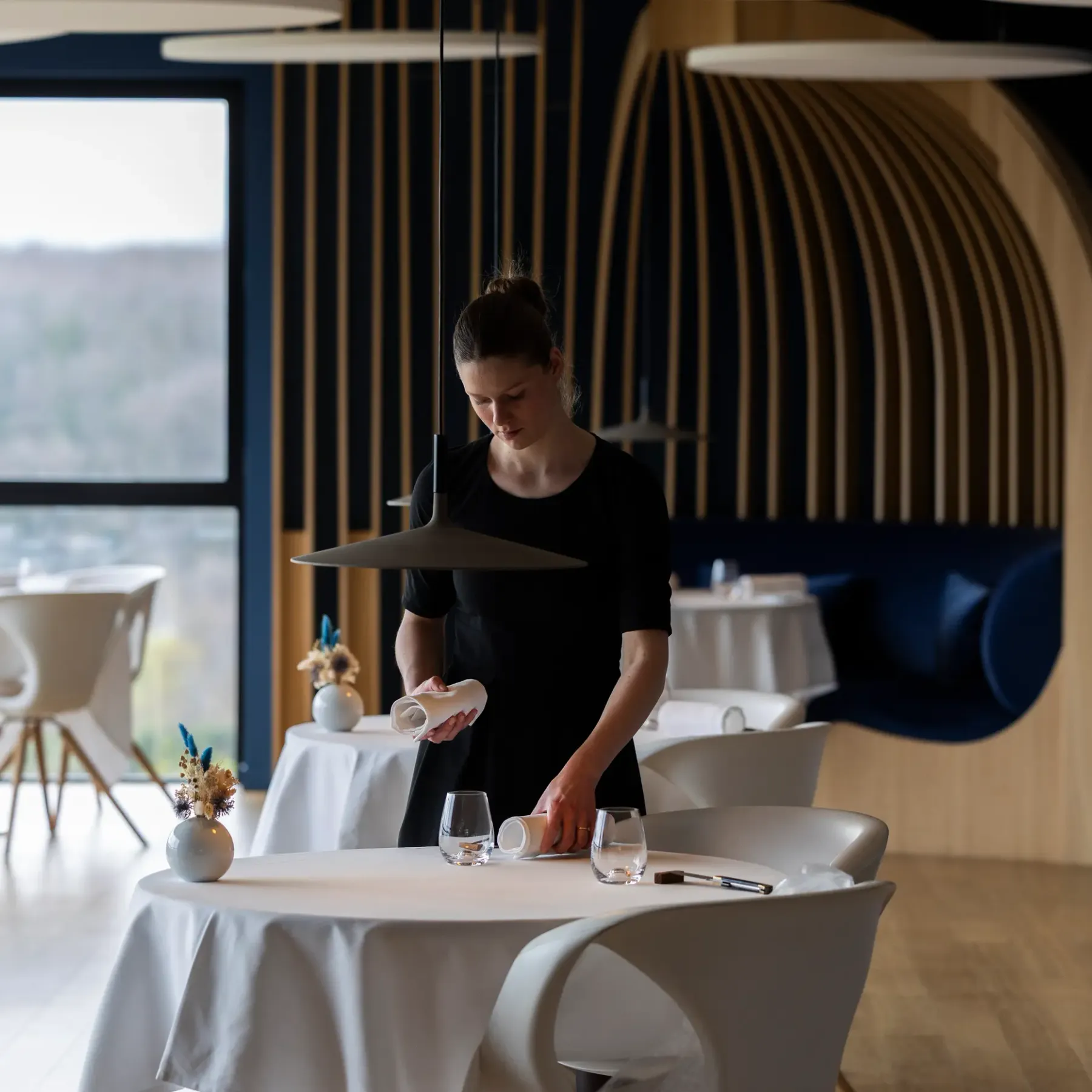 A person in black attire is preparing a table in a modern restaurant with large glass windows.