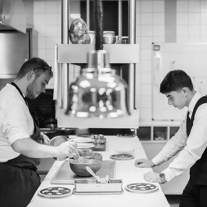 Two people in professional attire are preparing dishes in a kitchen equipped with restaurant equipment.