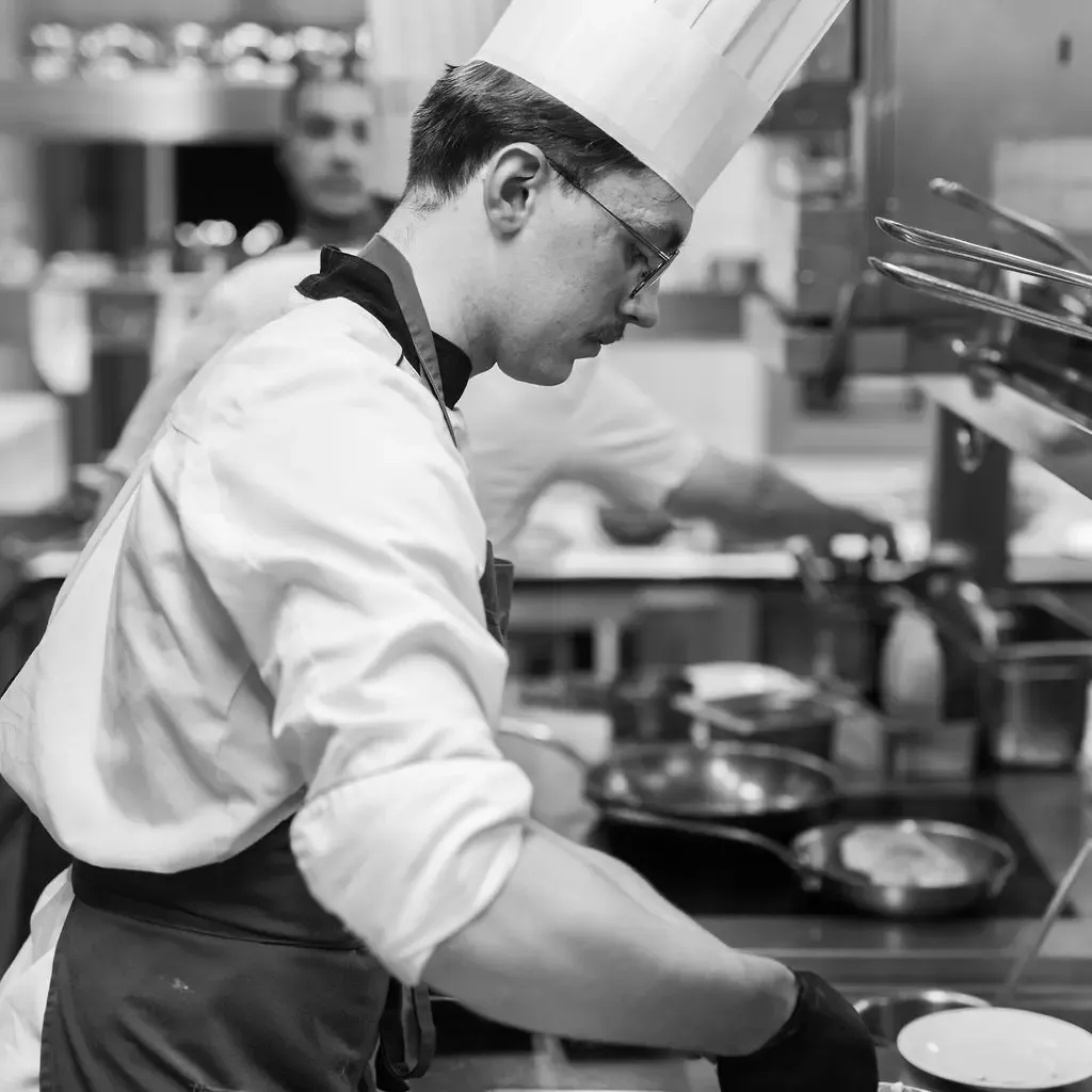A chef wearing a white toque works in a professional kitchen, focused on his culinary preparation.