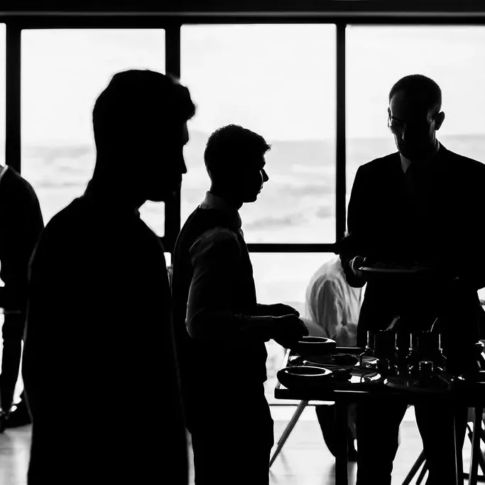 Four silhouetted figures discuss around a table with cups, in front of a large window offering an outdoor view.