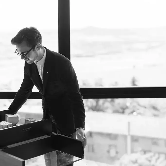 A man in a suit and glasses selects pastries from an open display on a table near a large window with a view of a snowy urban landscape.