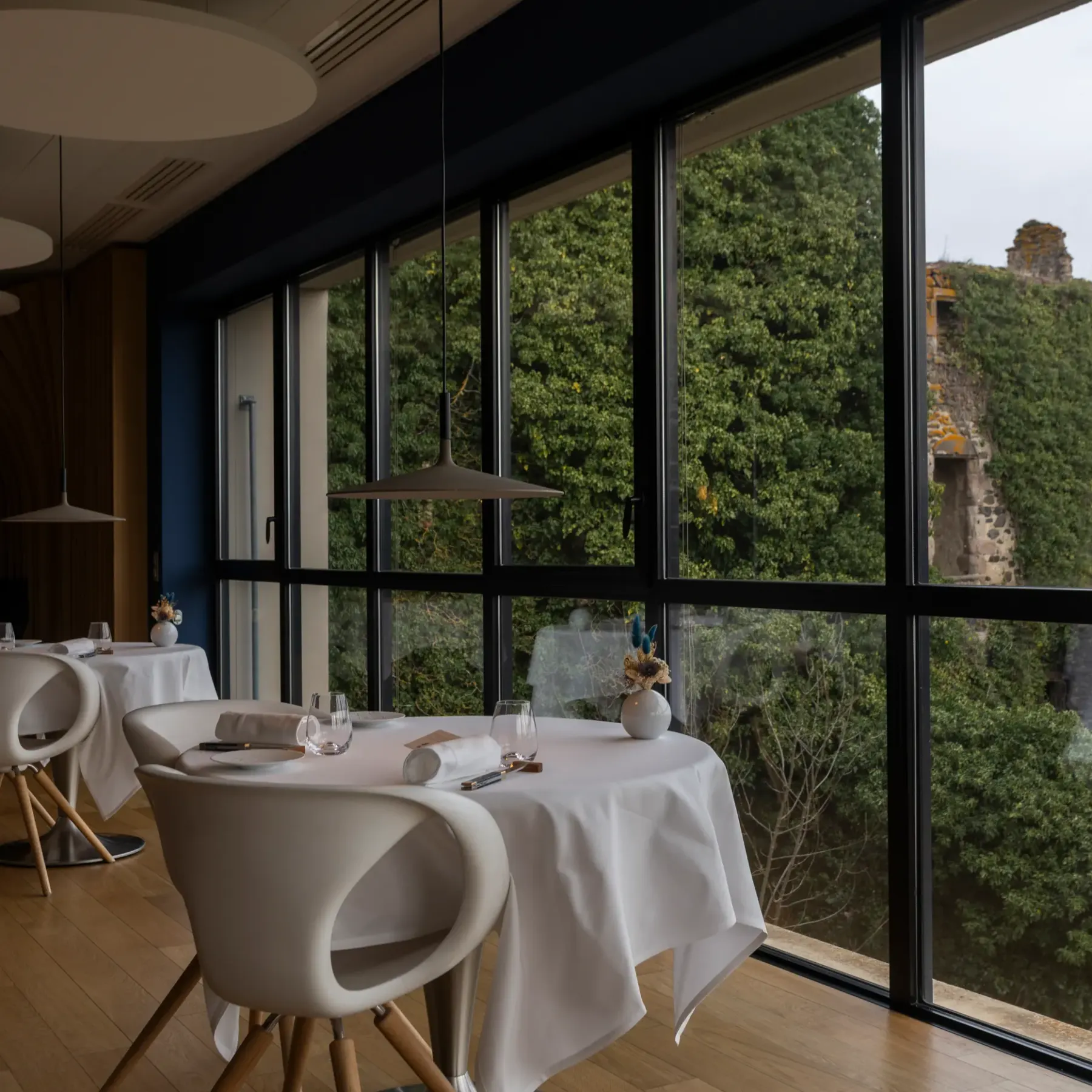 Restaurant with white tables and chairs in front of large windows offering a view of a wooded hill and ruins.