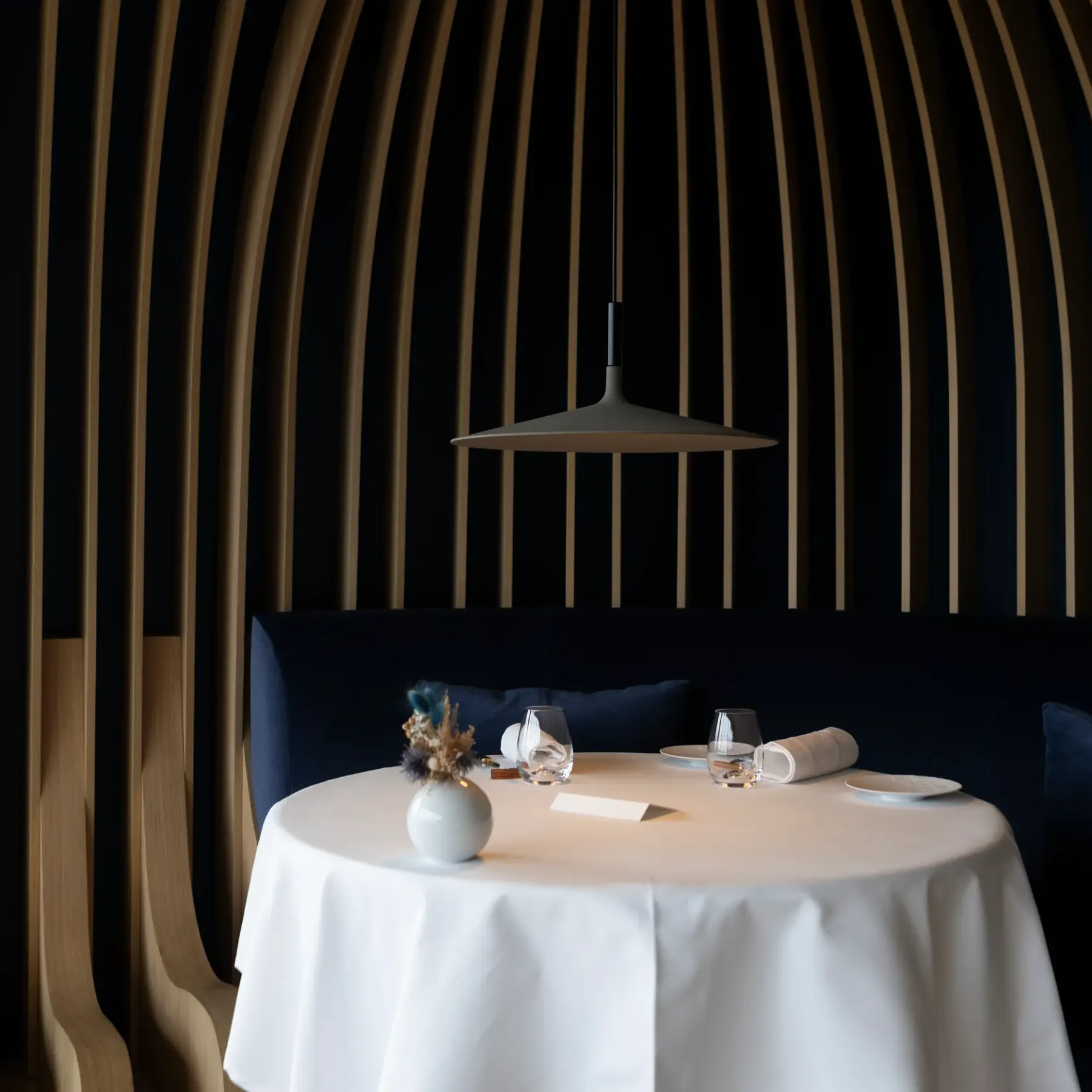 Table set with white tablecloth, flower vase, glasses, and rolled napkin, under a pendant light, in a space with dark and light vertical striped walls.