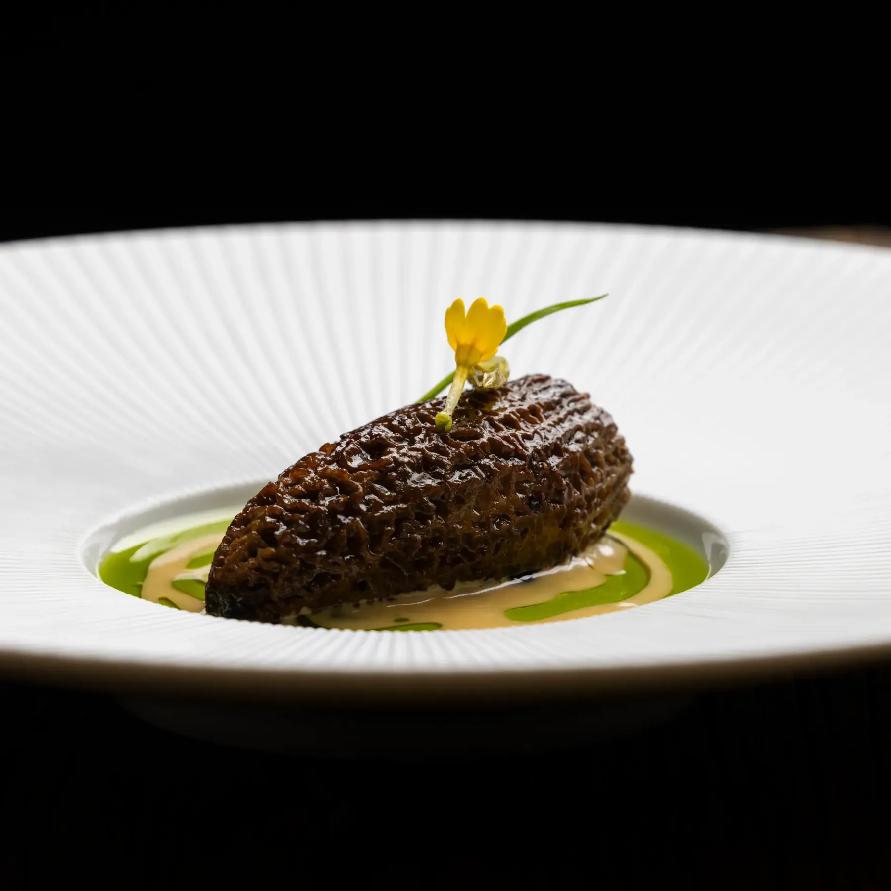 A dark brown piece of food is placed on a white plate with green sauce around it, accompanied by a small yellow flower.