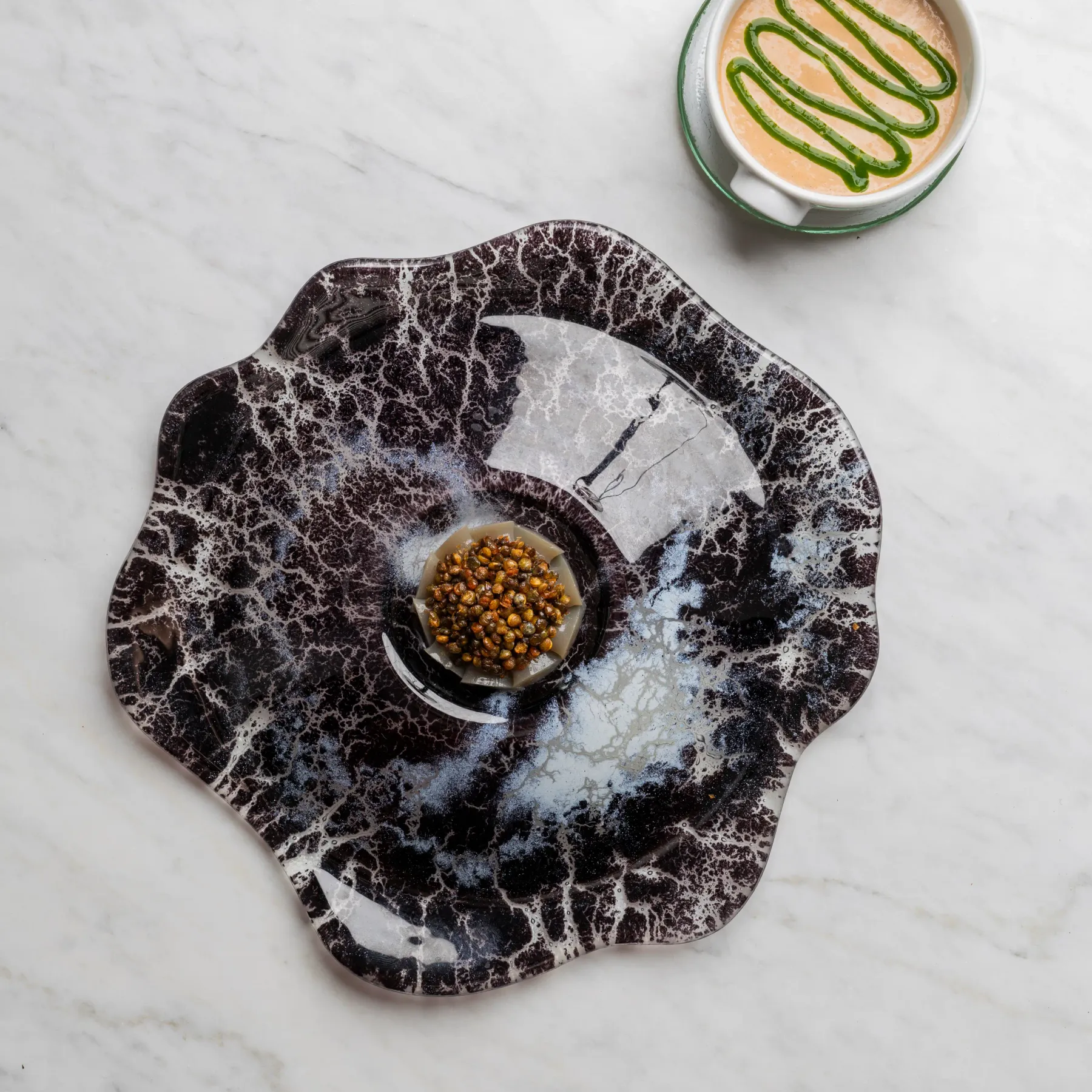 A cup of coffee with a green decoration on the surface is placed on a saucer, accompanied by a plate containing seeds on a white marble surface.