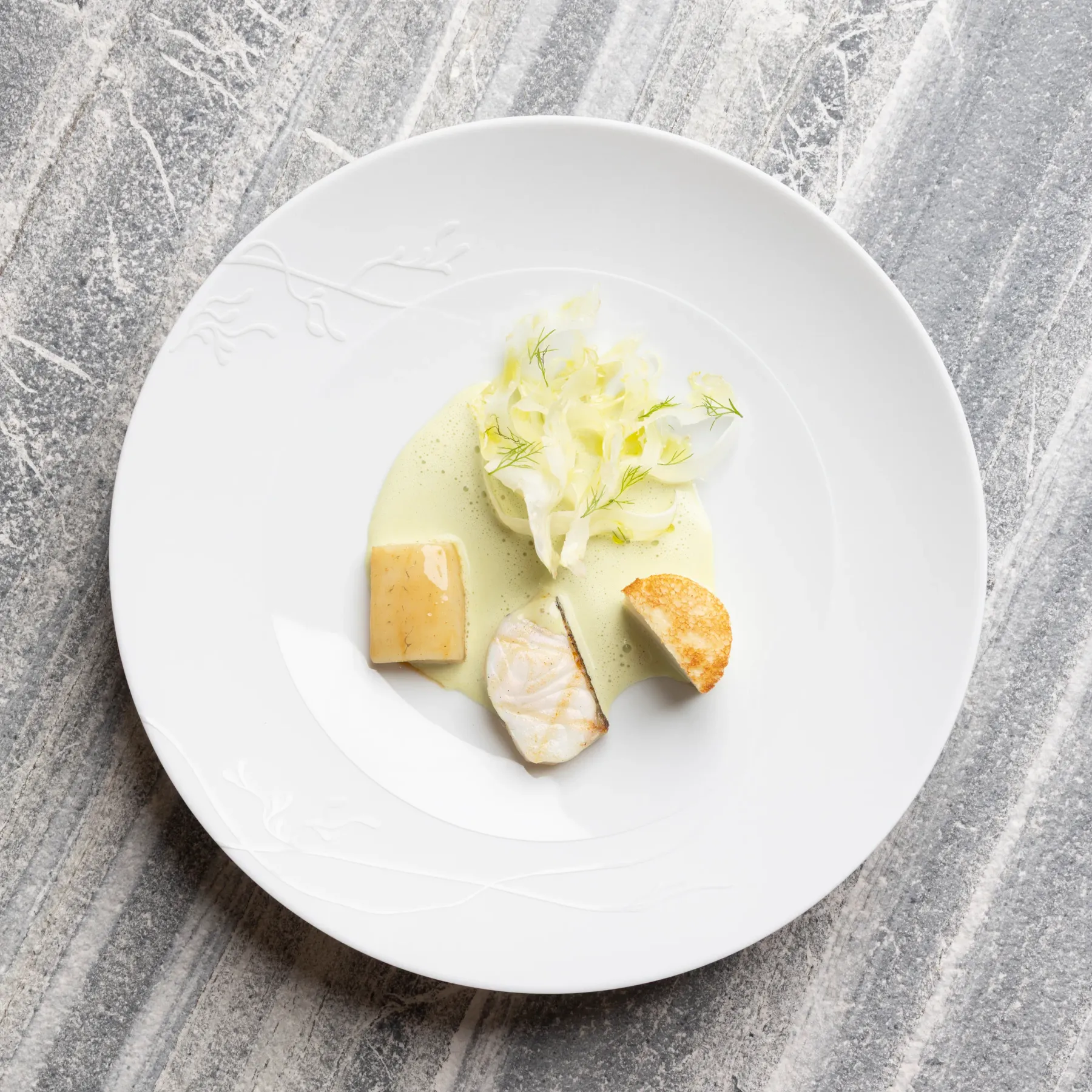 A dish with a green sauce, pieces of fish, vegetables, and croutons on a white plate placed on a gray marble surface.