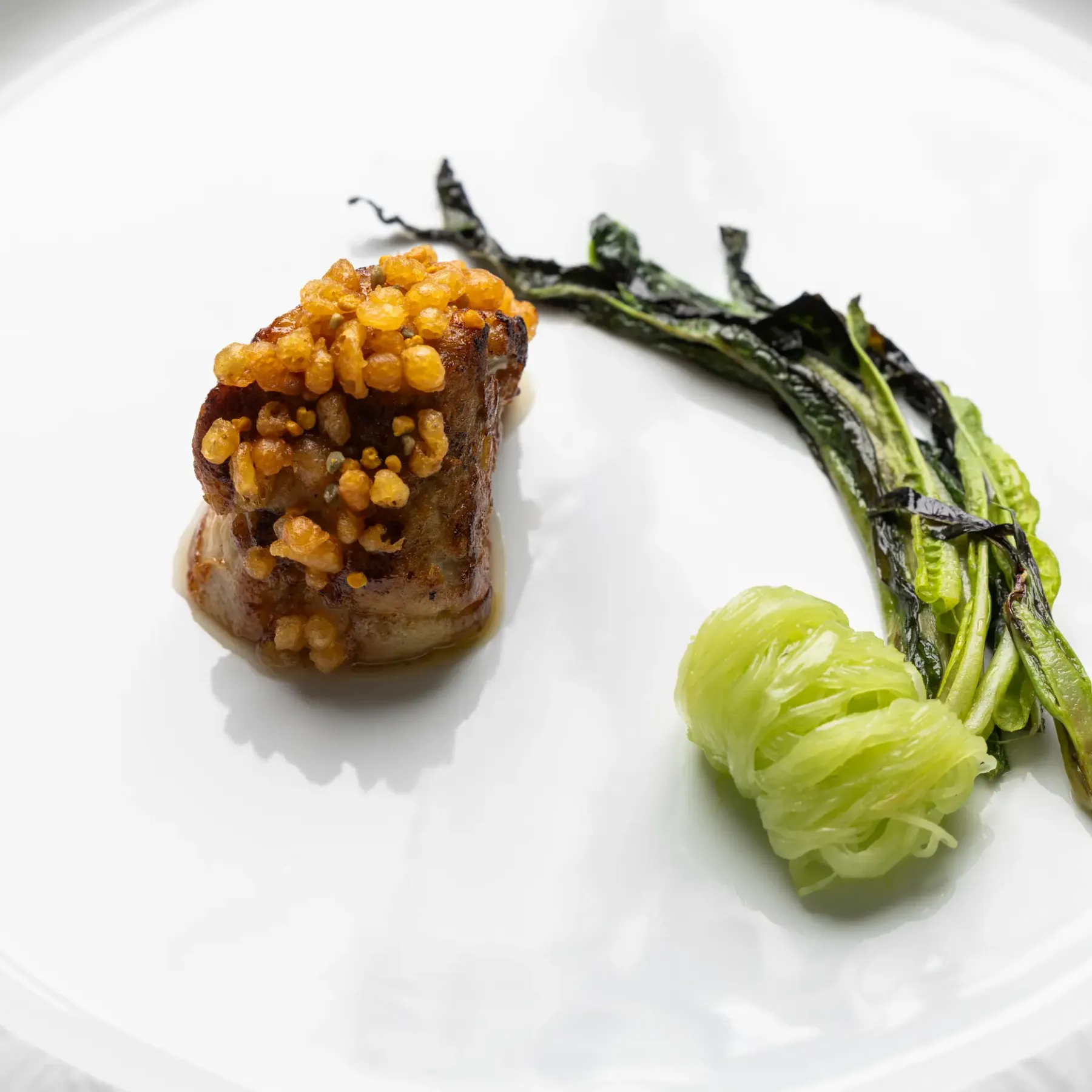 A piece of meat garnished with golden grains served with green vegetables and a translucent ball on a white plate.