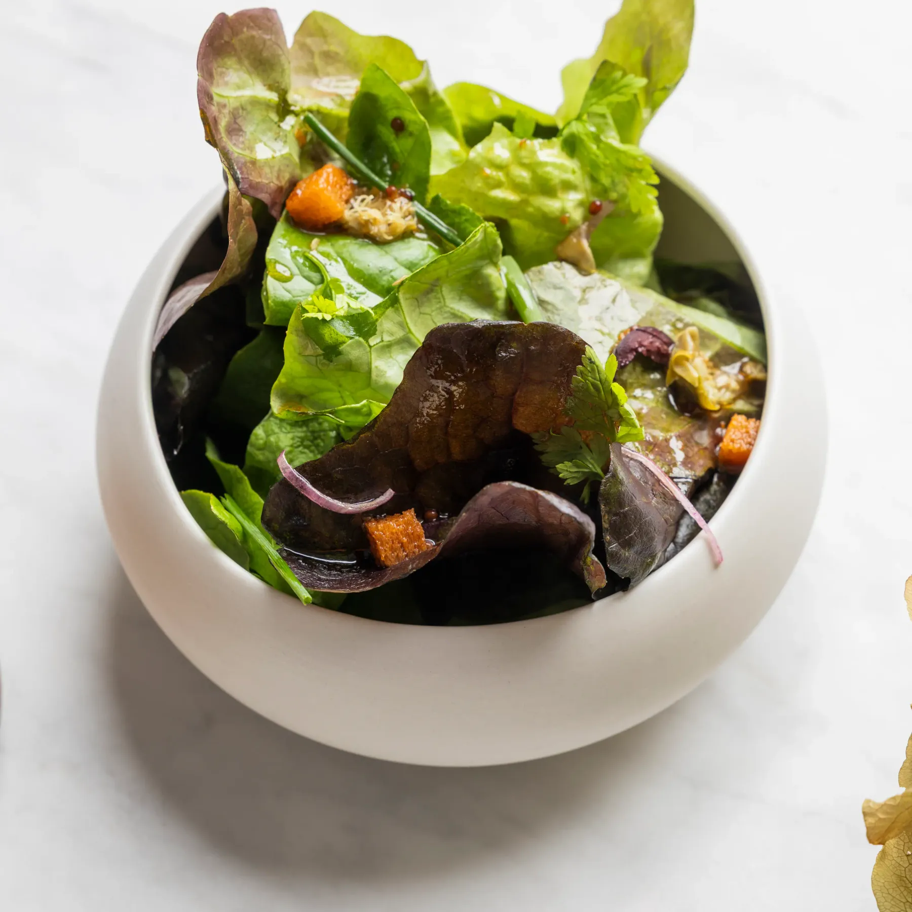 A white bowl contains a salad made of green leaves, red onions, roasted vegetable pieces, and croutons.