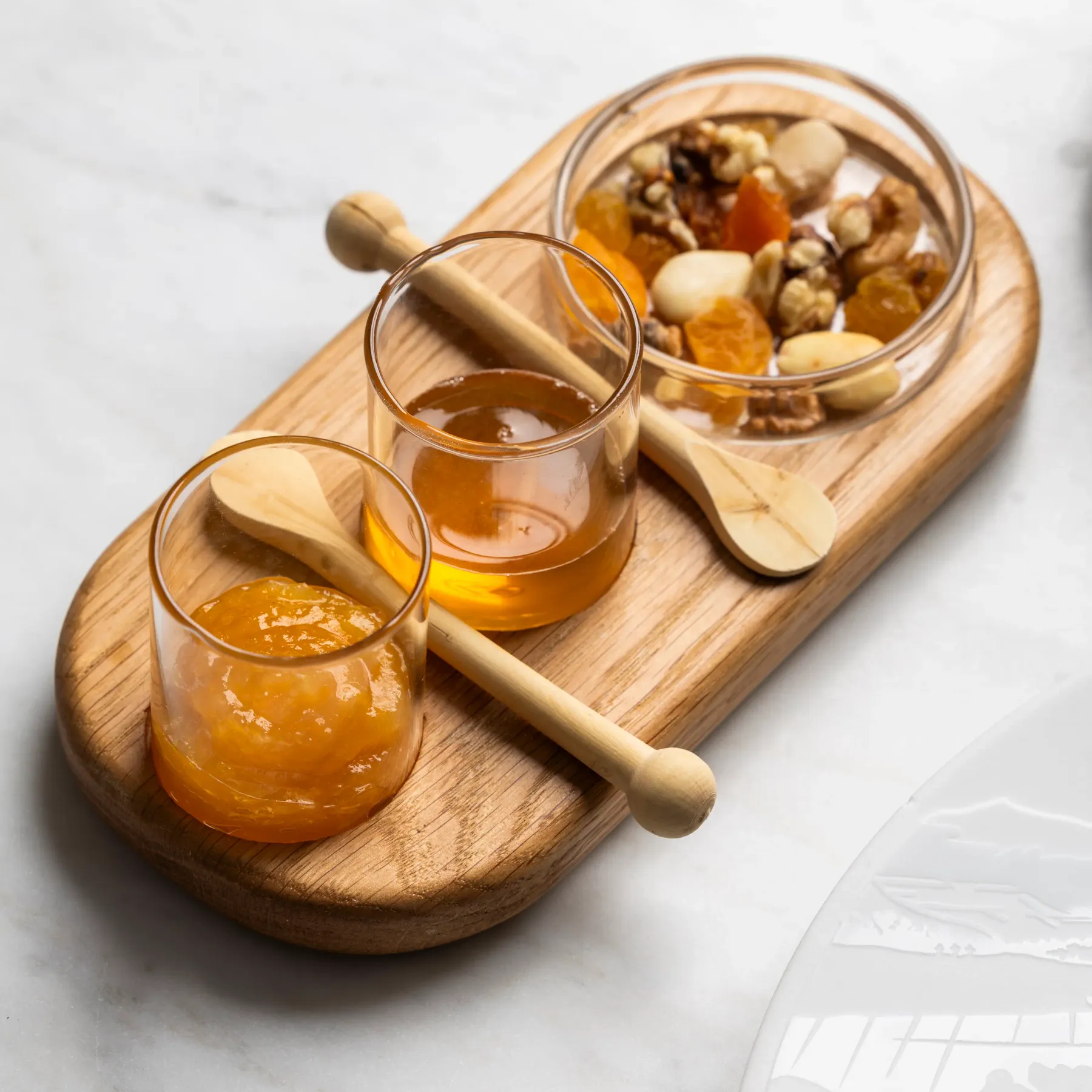 A wooden tray with small jam jars, honey glasses, and a bowl of nuts and dried fruit.